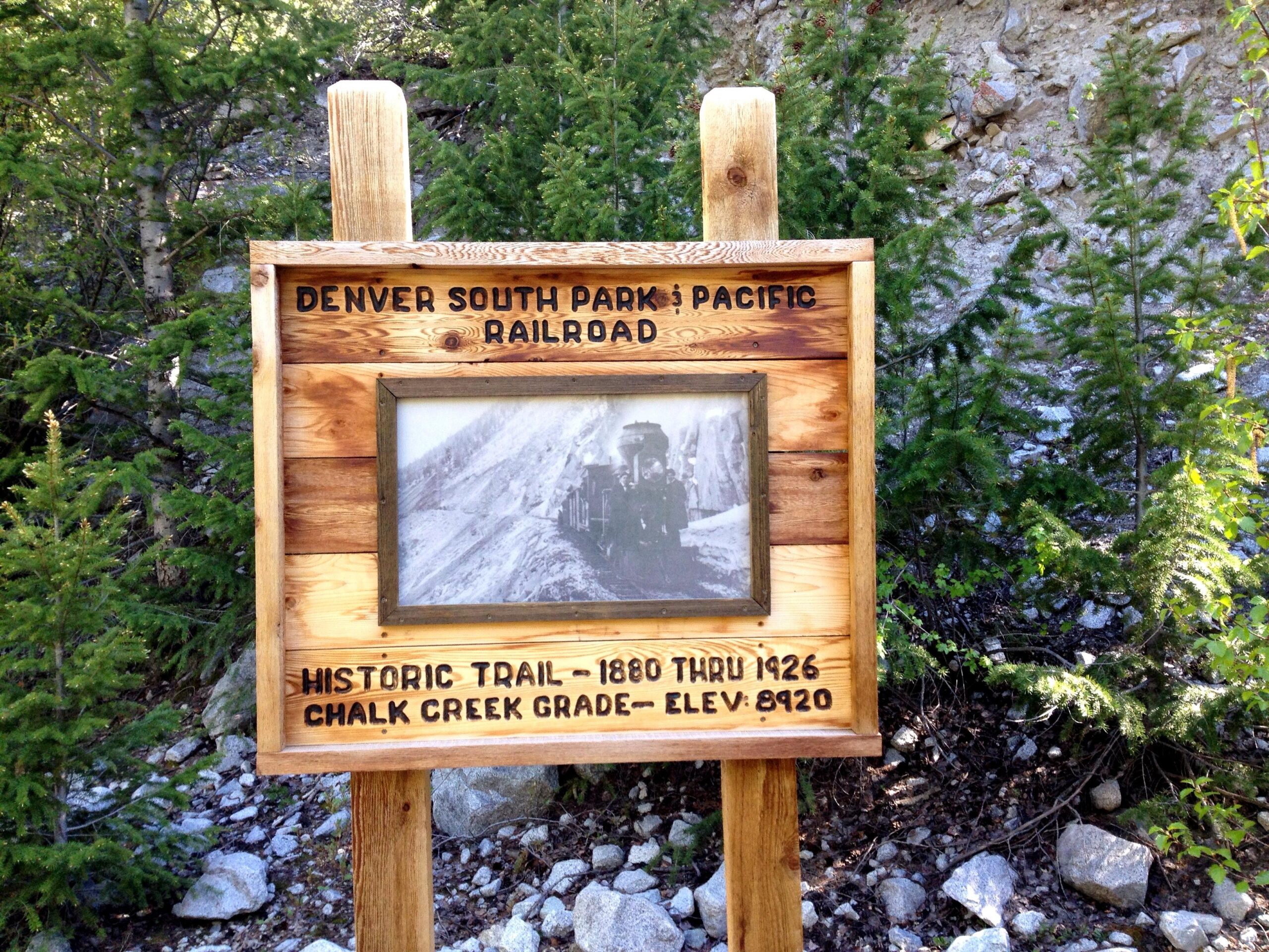 Wooden sign detailing the Denver South Park & Pacific Railroad, featuring a historic photo of a train. The sign includes text that reads "Historic Trail - 1880 thru 1926, Chalk Creek Grade - Elev: 8920" and is surrounded by greenery. Narrow Gauge Trail mountain bike trail.