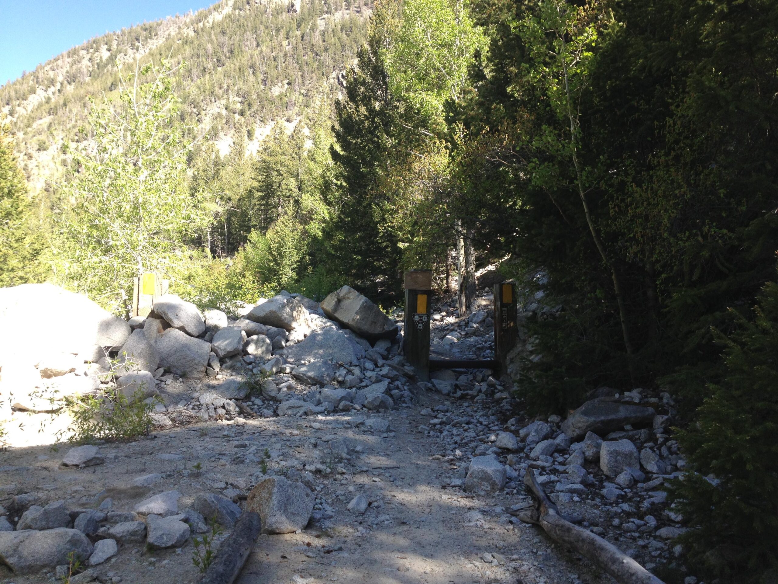 A rocky trail leading into a forested area, with large boulders scattered along the path and a small wooden entrance marked with signage on either side. Tall trees and a mountain slope are visible in the background, under a clear blue sky. Narrow Gauge Trail mountain bike trail.