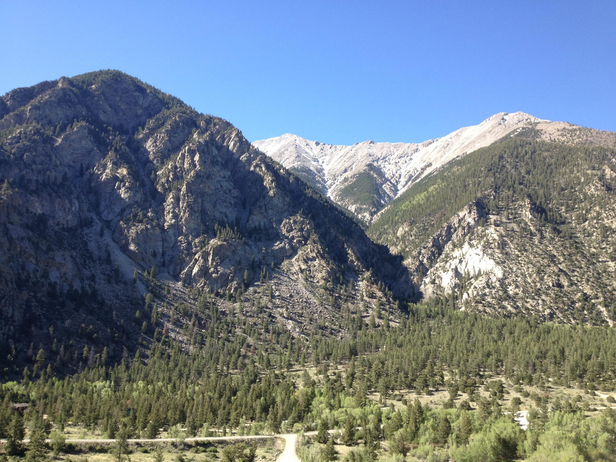 A panoramic view of rugged mountains with steep rocky slopes covered in patches of green pine trees against a clear blue sky. The foreground features a valley with a diverse tree landscape, while the peaks in the background show rocky surfaces, some dusted with snow. A winding road can be seen at the bottom of the image. Narrow Gauge Trail mountain bike trail.