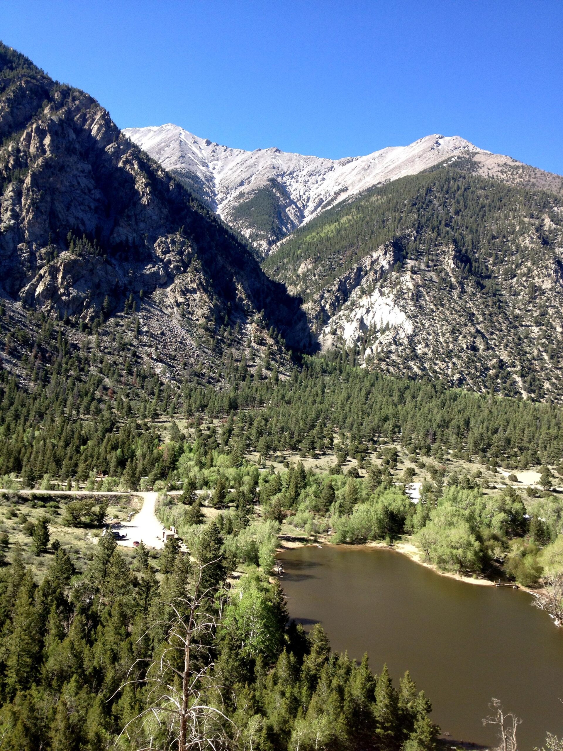 A scenic mountain landscape featuring rugged peaks with snow-capped summits in the background, surrounded by lush green forests. In the foreground, a calm pond reflects the surrounding greenery, with a dirt road and parking area visible near the water's edge. The sky is clear and blue, enhancing the natural beauty of the scene. Narrow Gauge Trail mountain bike trail.
