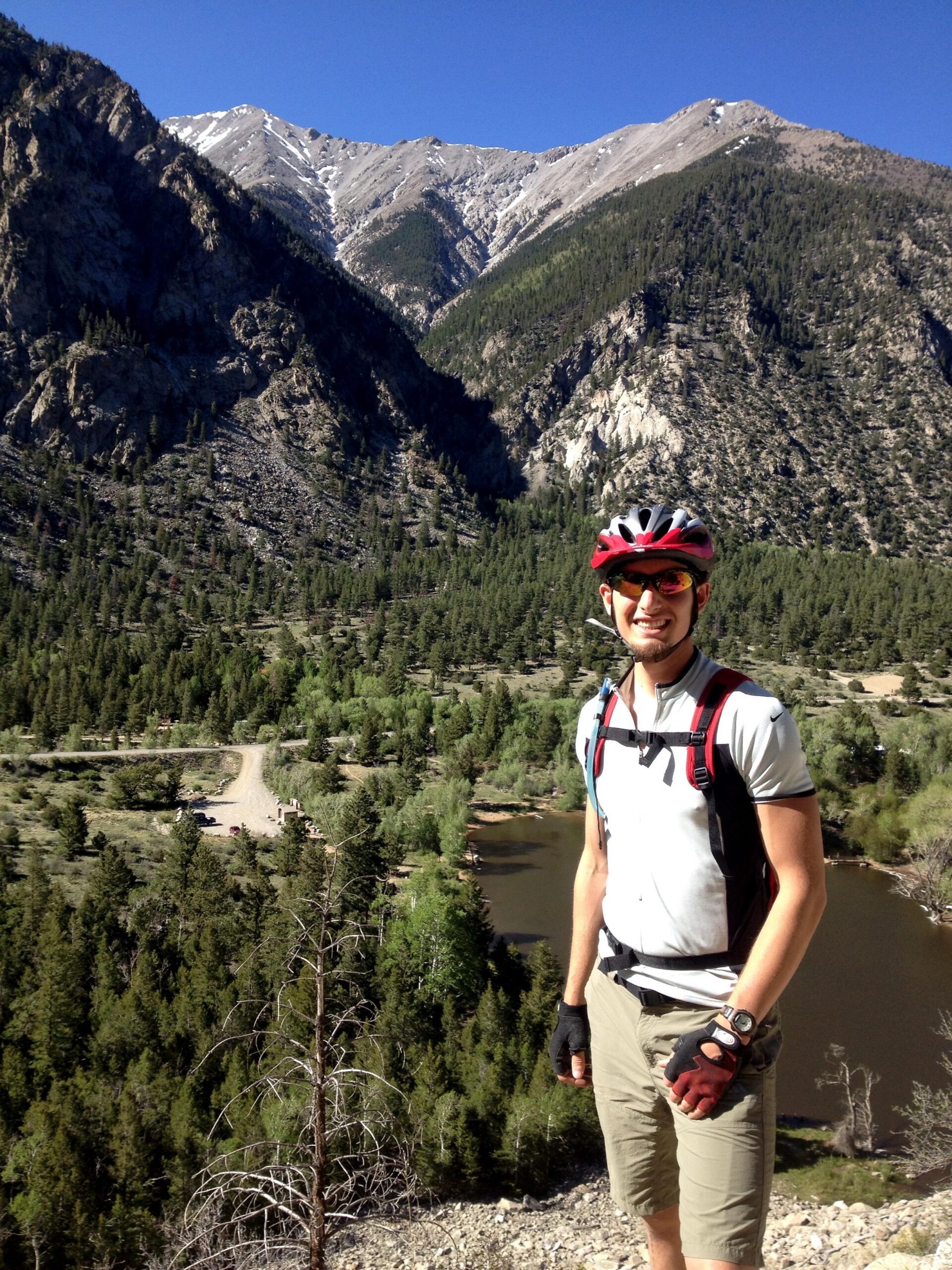 A cyclist wearing a helmet and sunglasses stands on a rocky outcrop with a scenic mountain backdrop. Lush greenery and a winding river are visible below, alongside a dirt road and parked vehicles in the distance. The sky is clear and blue, indicating a sunny day. Narrow Gauge Trail mountain bike trail.