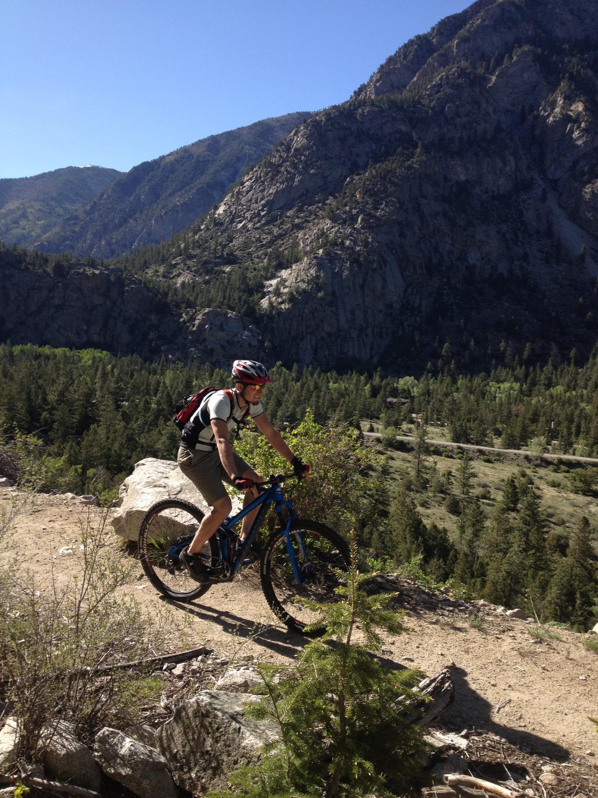 A mountain biker navigating a rocky trail in a mountainous landscape, surrounded by lush greenery and towering cliffs under a clear blue sky. Narrow Gauge Trail mountain bike trail.