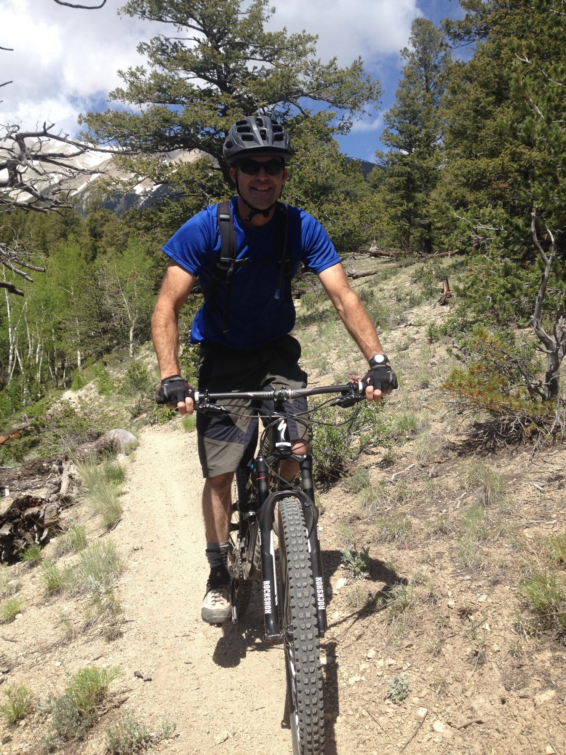 A person riding a mountain bike on a dirt trail surrounded by trees and greenery, wearing a black helmet and sunglasses. The background features mountains and a partly cloudy sky. The individual is smiling and appears to be enjoying the ride. Colorado Trail: Mount Princeton to Avalanche Trailhead / Collegiate Peaks Wilderness mountain bike trail.
