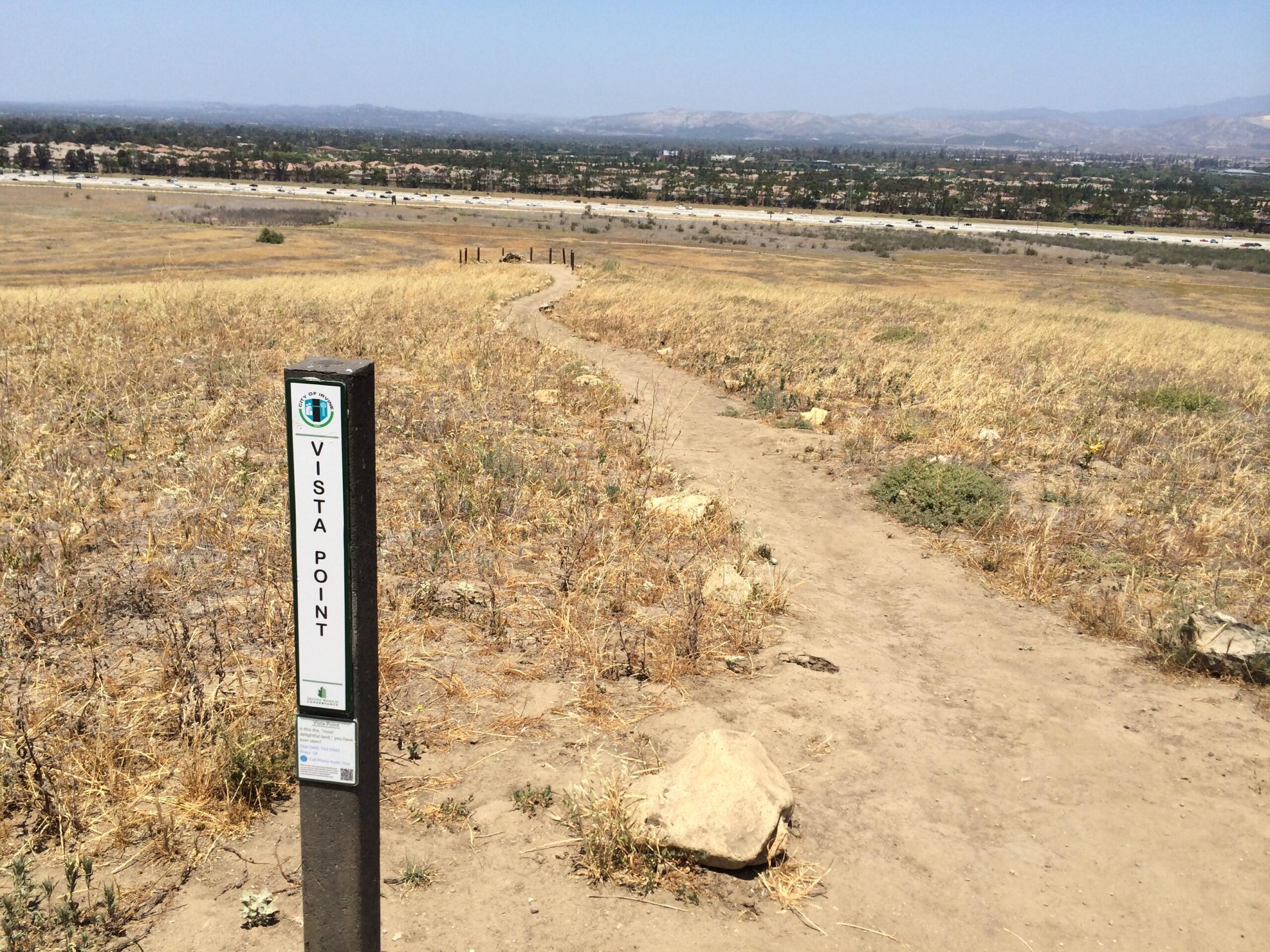A dirt path leading to a vista point sign, with a view of a valley in the background. The landscape features dry grasses and a distant highway lined with trees and buildings. Clear blue sky overhead. Quail Hill Loop mountain bike trail.