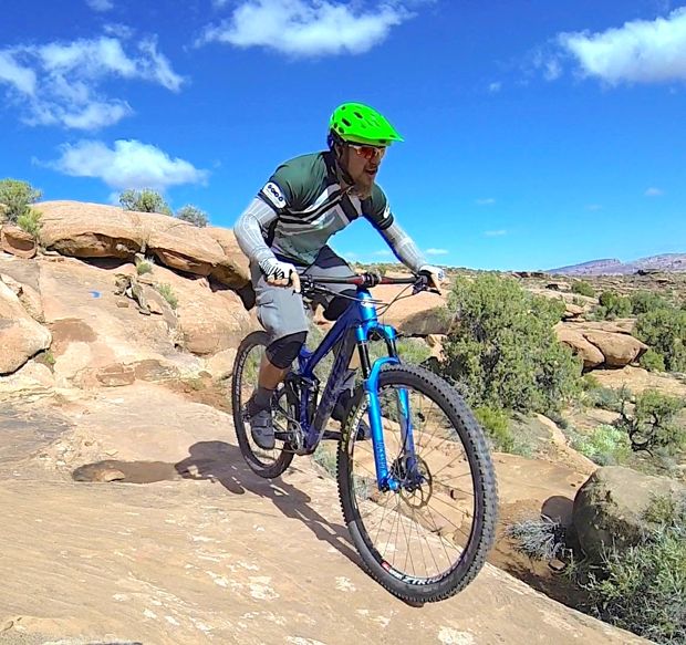 A cyclist navigating rocky terrain on a mountain bike, wearing a green helmet and gray sports attire, under a bright blue sky with a few clouds. Surrounding the cyclist are scattered rocks and patches of greenery.
