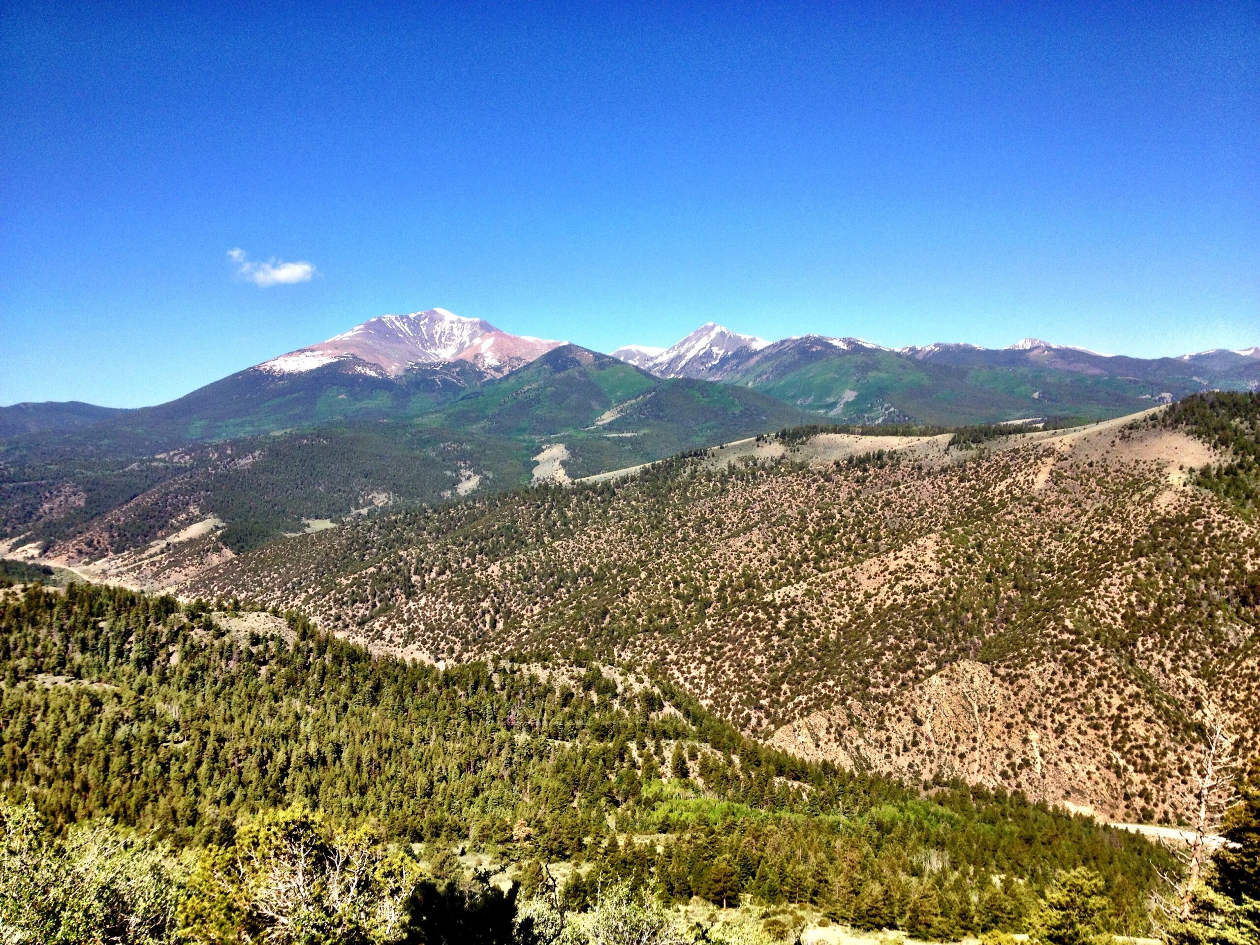 A panoramic view of a mountainous landscape under a clear blue sky. The scene features a range of peaks, some capped with snow, surrounded by lush green forests and rolling hills. The foreground showcases a variety of tree-covered slopes, creating a serene and picturesque natural setting. Rainbow Trail: Hwy 285 to Methodist Mountain Thd mountain bike trail.