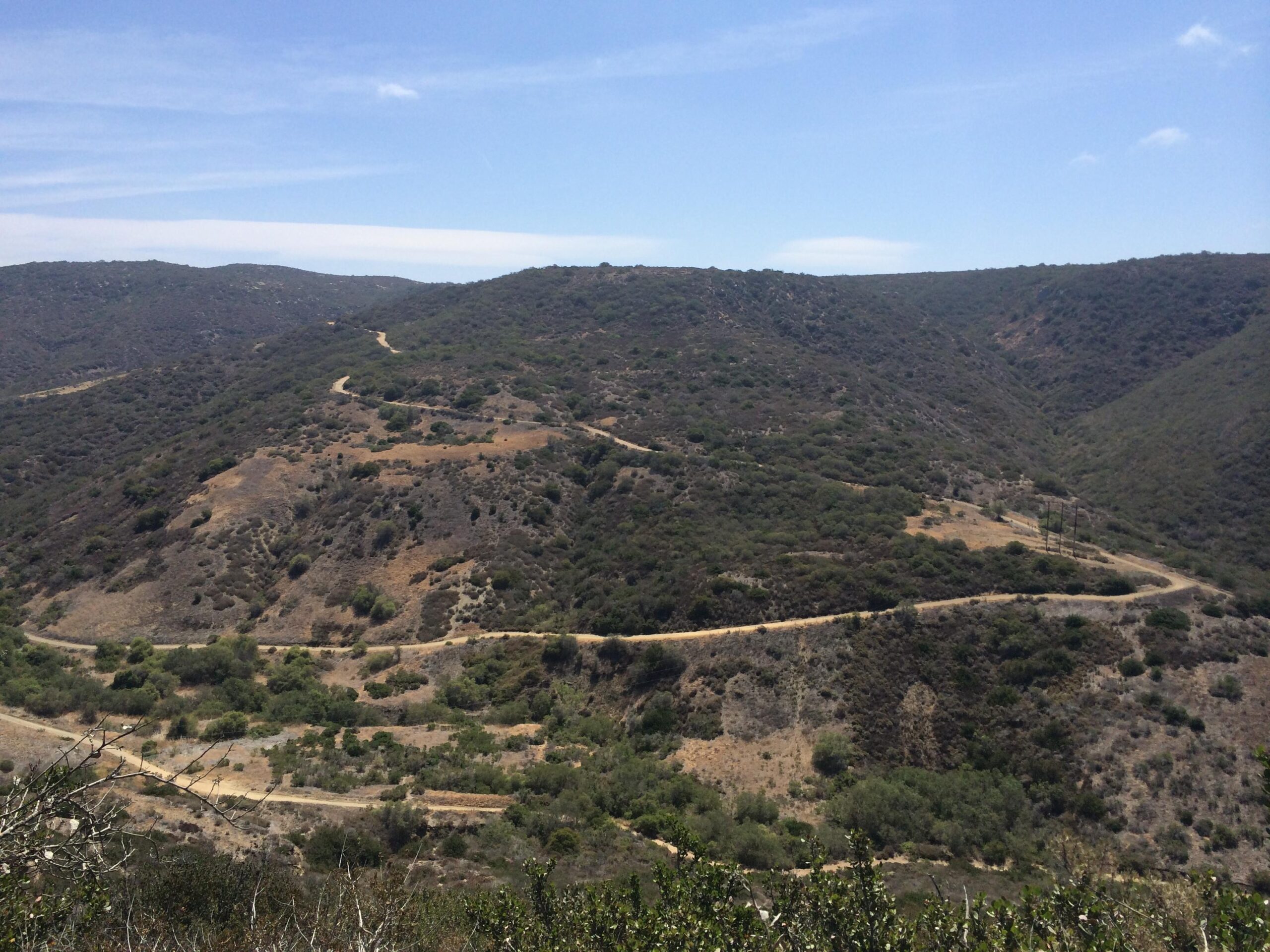 A panoramic view of rolling hills covered with shrubs and sparse vegetation, with winding dirt trails intersecting the landscape under a clear blue sky. East Cut Across mountain bike trail.