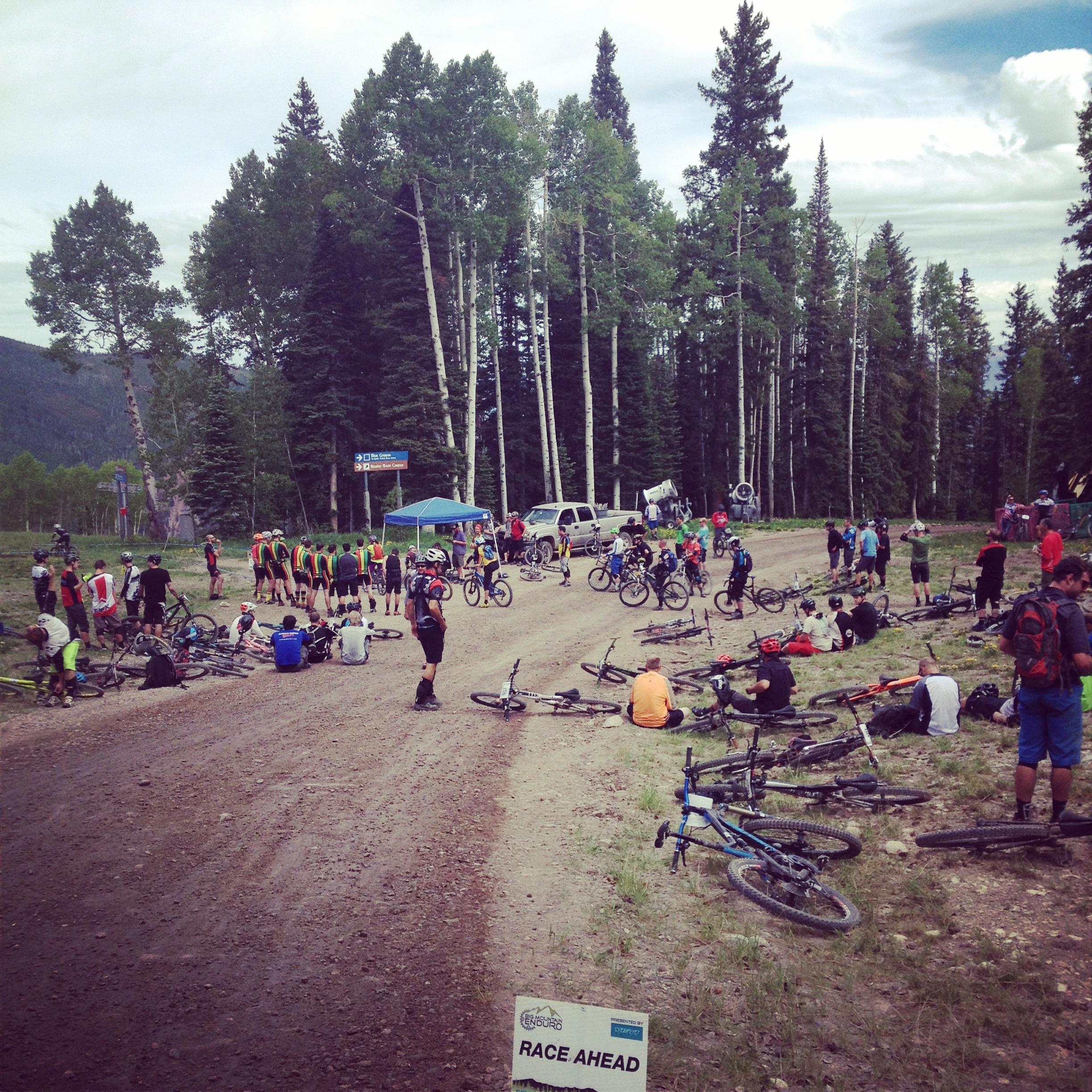 A group of mountain bikers gather at a race start point on a dirt road surrounded by tall trees. Some participants are on their bikes, while others are sitting or standing nearby. A sign reading "Race Ahead" is visible in the foreground, and a tent with people is set up in the background. The atmosphere appears lively and active, indicative of a biking event. Village Bound mountain bike trail.