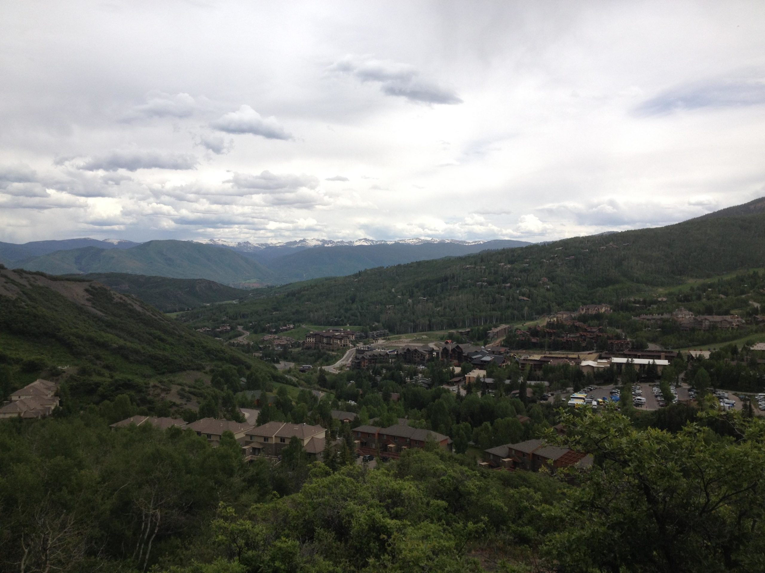 A panoramic view of a valley surrounded by mountains, featuring green hills, scattered residential buildings in the foreground, and a town visible in the middle ground. The sky is mostly cloudy, with patches of blue peeking through, and snow-capped peaks visible in the distance. Rim Trail mountain bike trail.