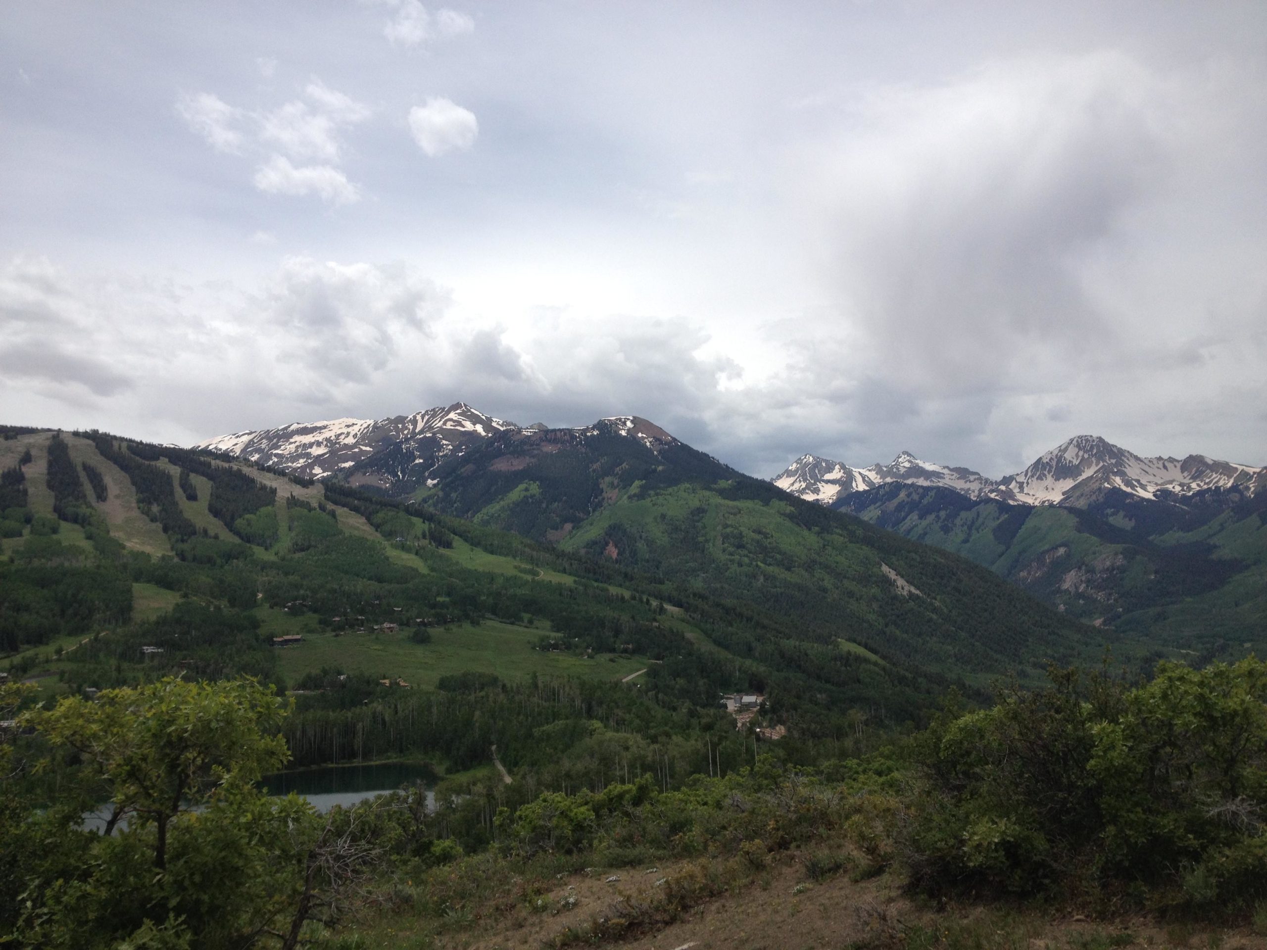 A panoramic view of lush green mountains with patches of snow on the peaks under a cloudy sky. In the foreground, a serene lake is visible, surrounded by greenery and trees, while the mountains in the background rise majestically, showcasing a mix of rocky terrain and vegetation. Rim Trail mountain bike trail.