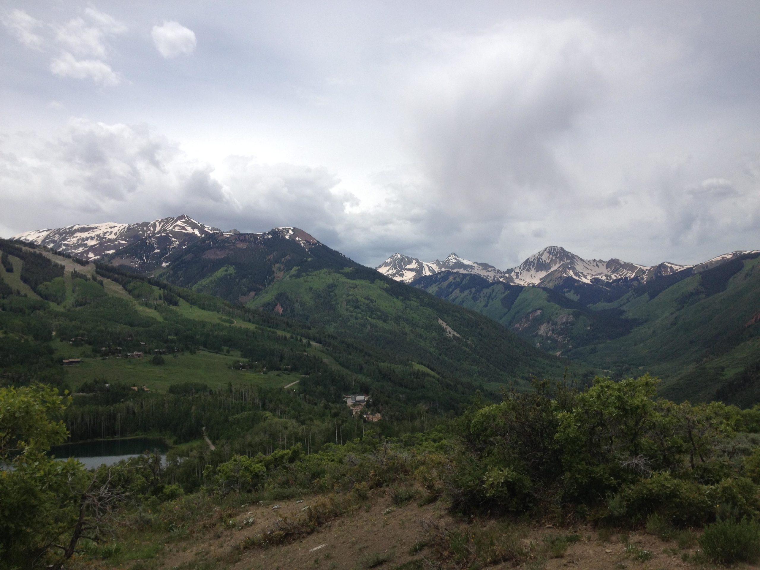 A panoramic view of lush green mountains under a cloudy sky, with snow-capped peaks rising in the background. A serene lake is visible among the trees at the lower part of the landscape, displaying a vibrant natural setting. Rim Trail mountain bike trail.