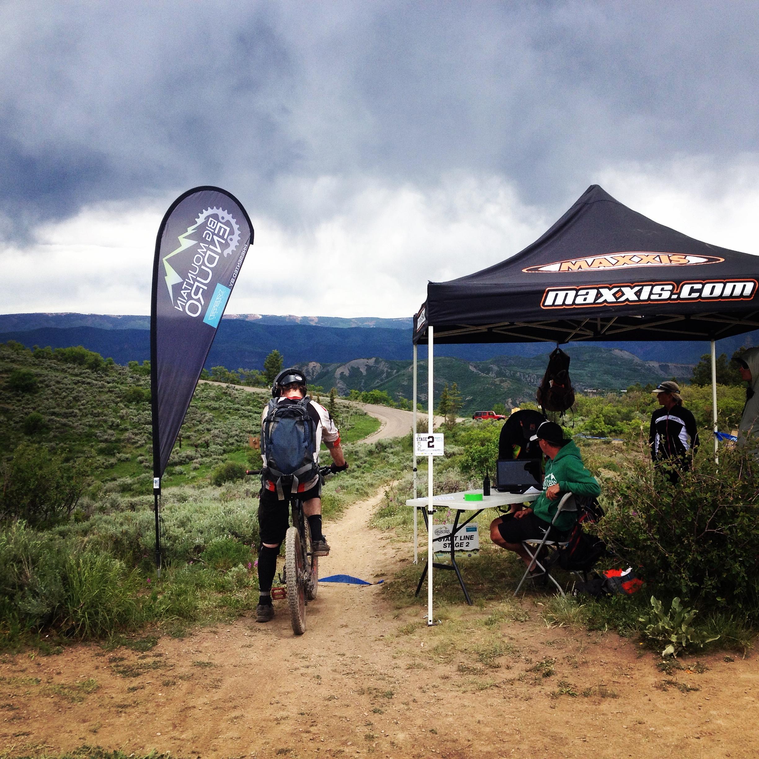A mountain biker approaches a small event tent set up alongside a dirt trail, with a flag featuring a logo in the background. People are gathered near the tent, which has a table displaying equipment and check-in materials. The sky is overcast, and green hills can be seen in the distance. Rim Trail mountain bike trail.