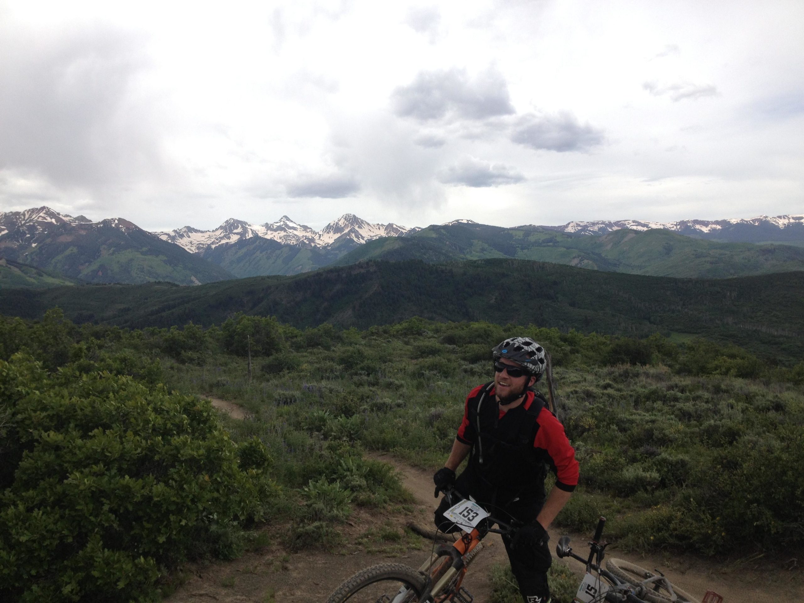 A mountain biker in protective gear stands on a trail, smiling and looking towards a picturesque mountain landscape featuring snow-capped peaks and lush green valleys under a partly cloudy sky. Two bicycles are positioned nearby on the ground. Rim Trail mountain bike trail.