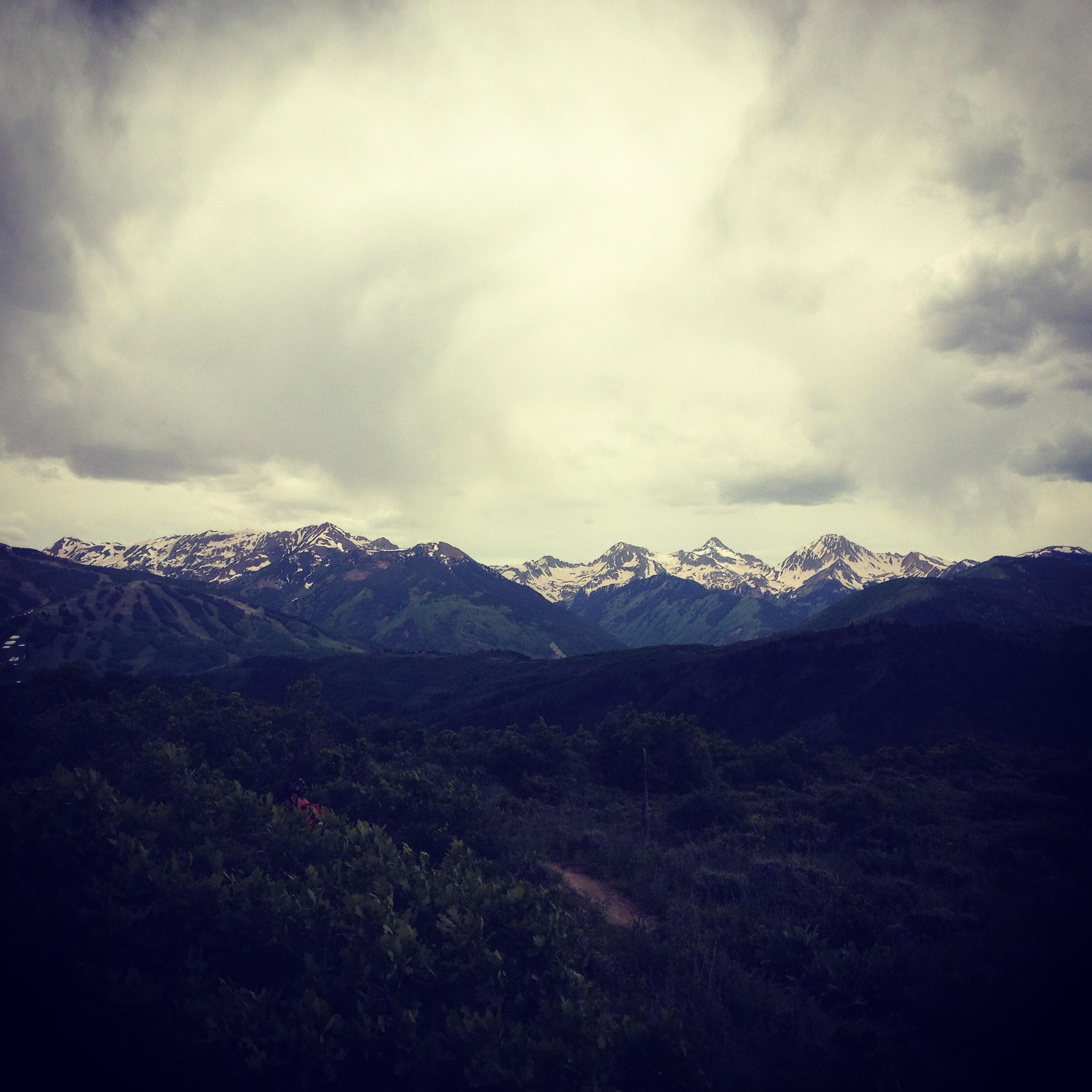 A panoramic view of snow-capped mountains under a cloudy sky, with lush green hills in the foreground. The landscape features a mix of rocky terrain and vegetation, suggesting a natural, serene environment. Rim Trail mountain bike trail.