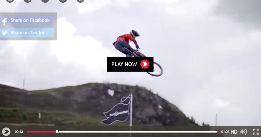 A mountain biker performing a jump against a cloudy sky, with a flag visible in the background. Social media sharing buttons for Facebook and Twitter are located at the top left, along with a "Play Now" button in the center. A video player timeline is displayed at the bottom.