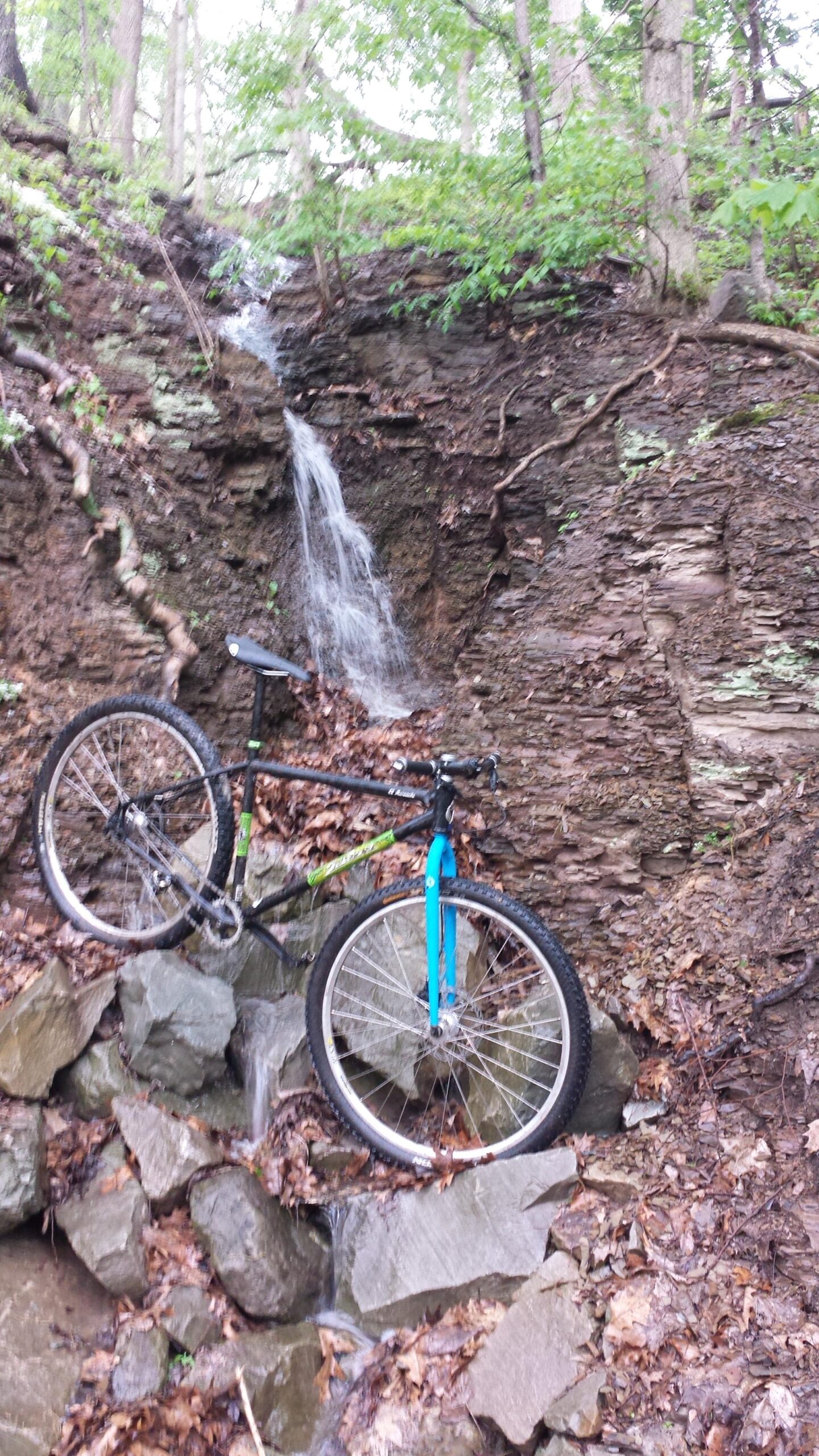 Salsa El Mariachi 3: A mountain bike leaning against a rocky terrain beside a small waterfall, surrounded by green foliage and fallen leaves.