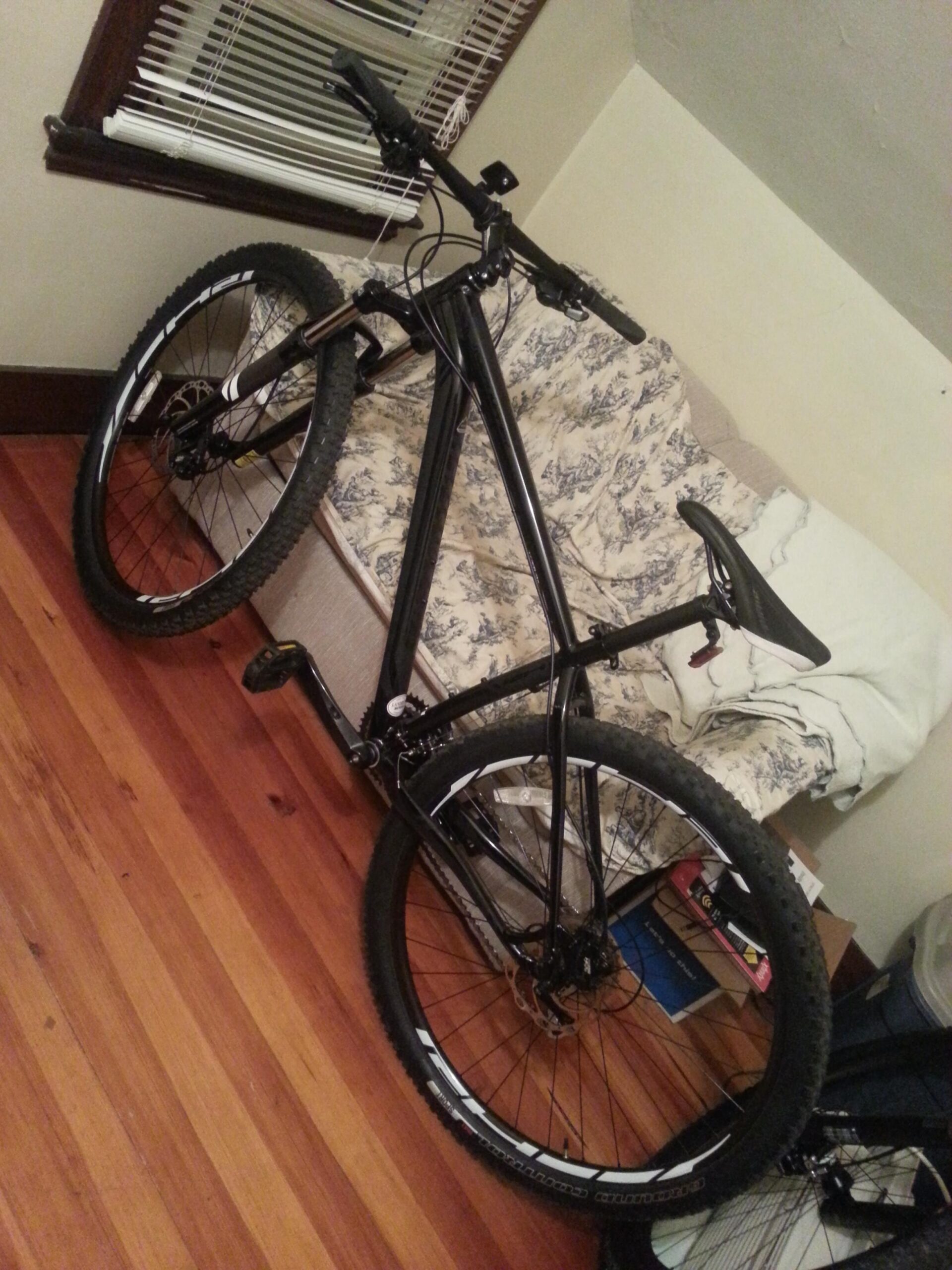 Specialized Rockhopper Comp 29: A black mountain bike is leaning against a couch in a room with wooden flooring. The bike features thick tires and a straightforward design, with the handlebars turned towards the viewer. In the background, there is a partially visible couch covered with a light-colored blanket, along with some boxes and a blue object on the floor. Natural light filters in through a window with blinds.