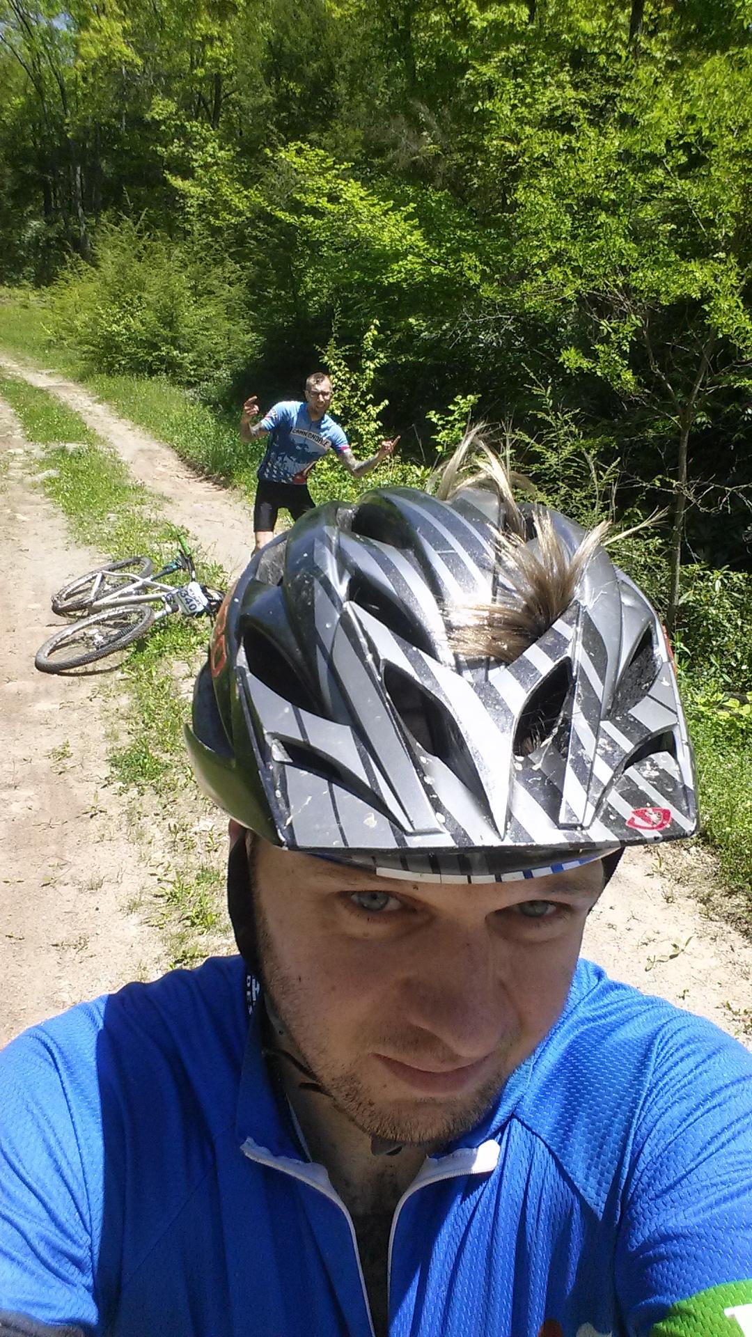 A person wearing a black and white striped bicycle helmet and a blue shirt takes a selfie on a dirt path surrounded by greenery. In the background, another individual with colorful cycling attire appears to be signaling or reacting to a situation. Two mountain bikes are lying on the ground nearby. Big Bear Lake Trail Center mountain bike trail.