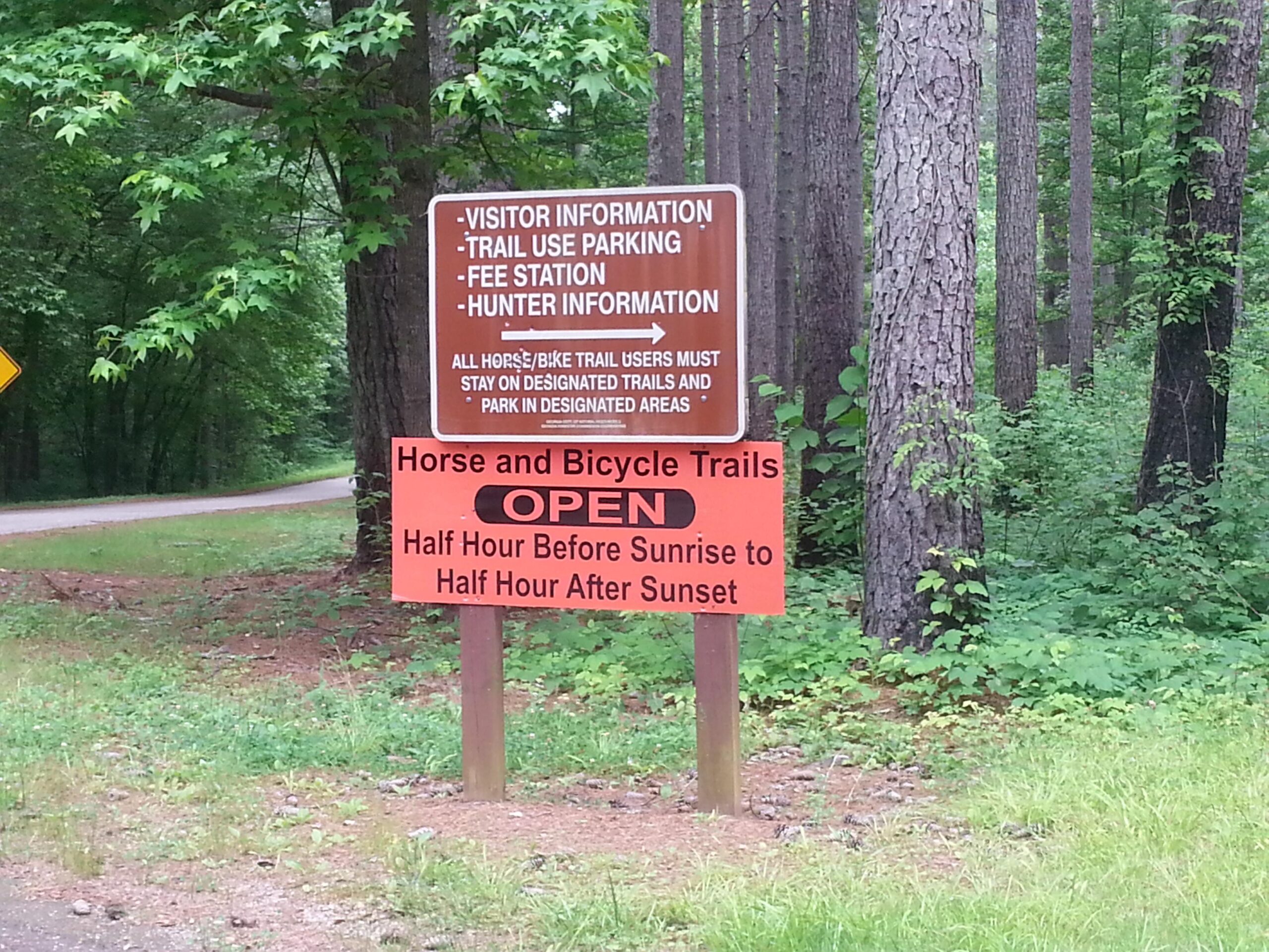 A wooden sign displaying information for visitors, including guidelines for trail use, park fees, and hunting information. The sign indicates that horse and bicycle trails are open from half an hour before sunrise to half an hour after sunset. The background features a wooded area with trees and greenery. Dawson Forest mountain bike trail.