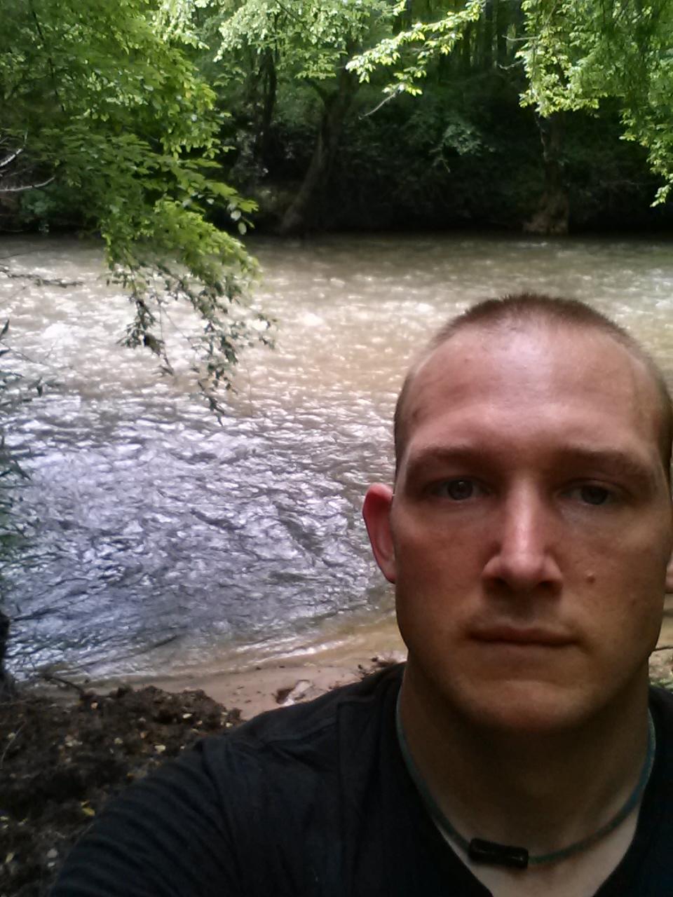 A close-up selfie of a person standing by a flowing creek surrounded by lush green trees. The water is slightly murky, and the sandy bank can be seen in the background. The individual has short hair and a serious expression, wearing a black shirt. Dawson Forest mountain bike trail.
