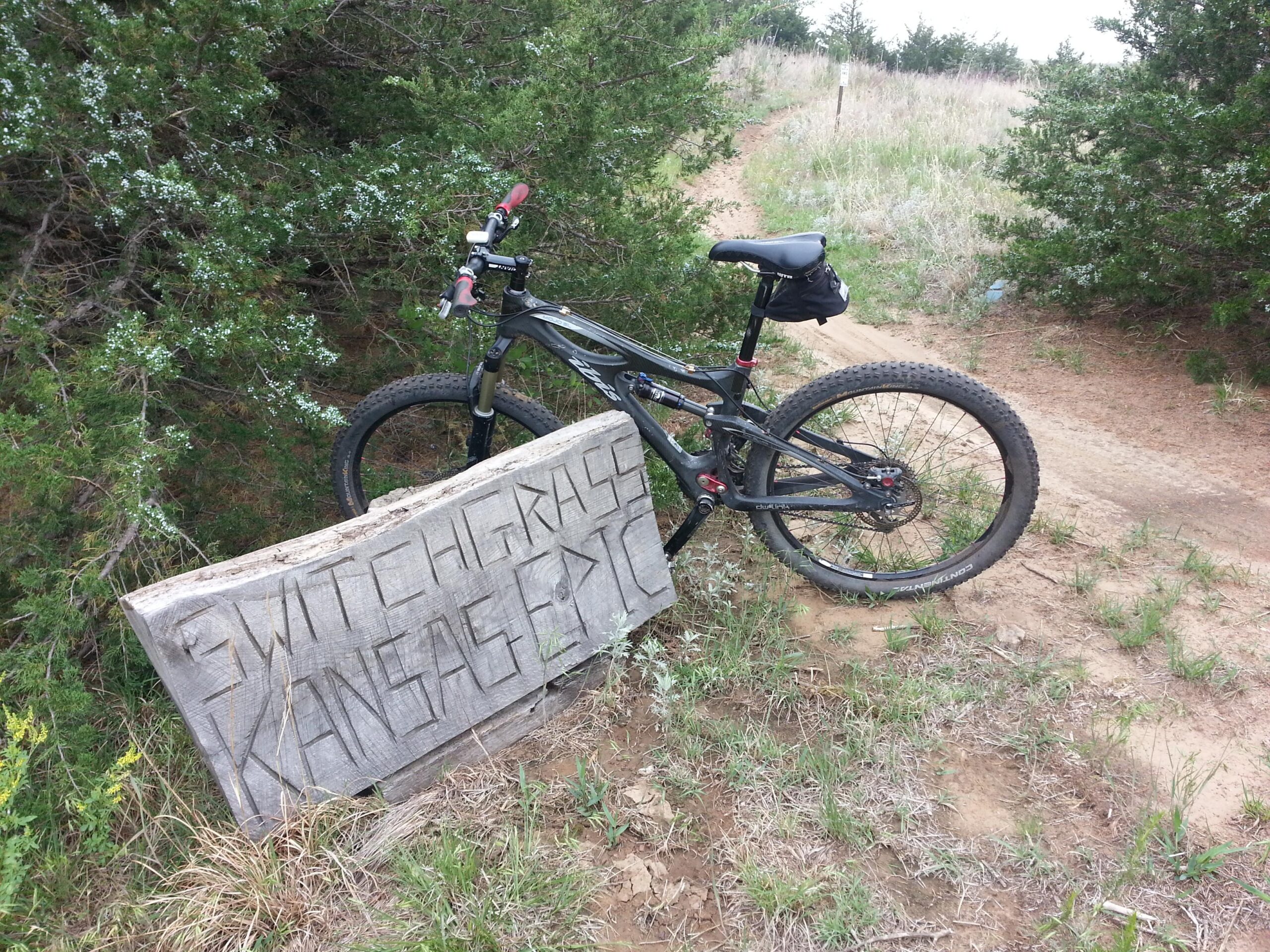 A black mountain bike resting against a wooden sign that reads "Switchgrass Kansas". The bike is positioned on a dirt path surrounded by tall grass and bushes. The scene depicts a natural outdoor setting. Switchgrass mountain bike trail.