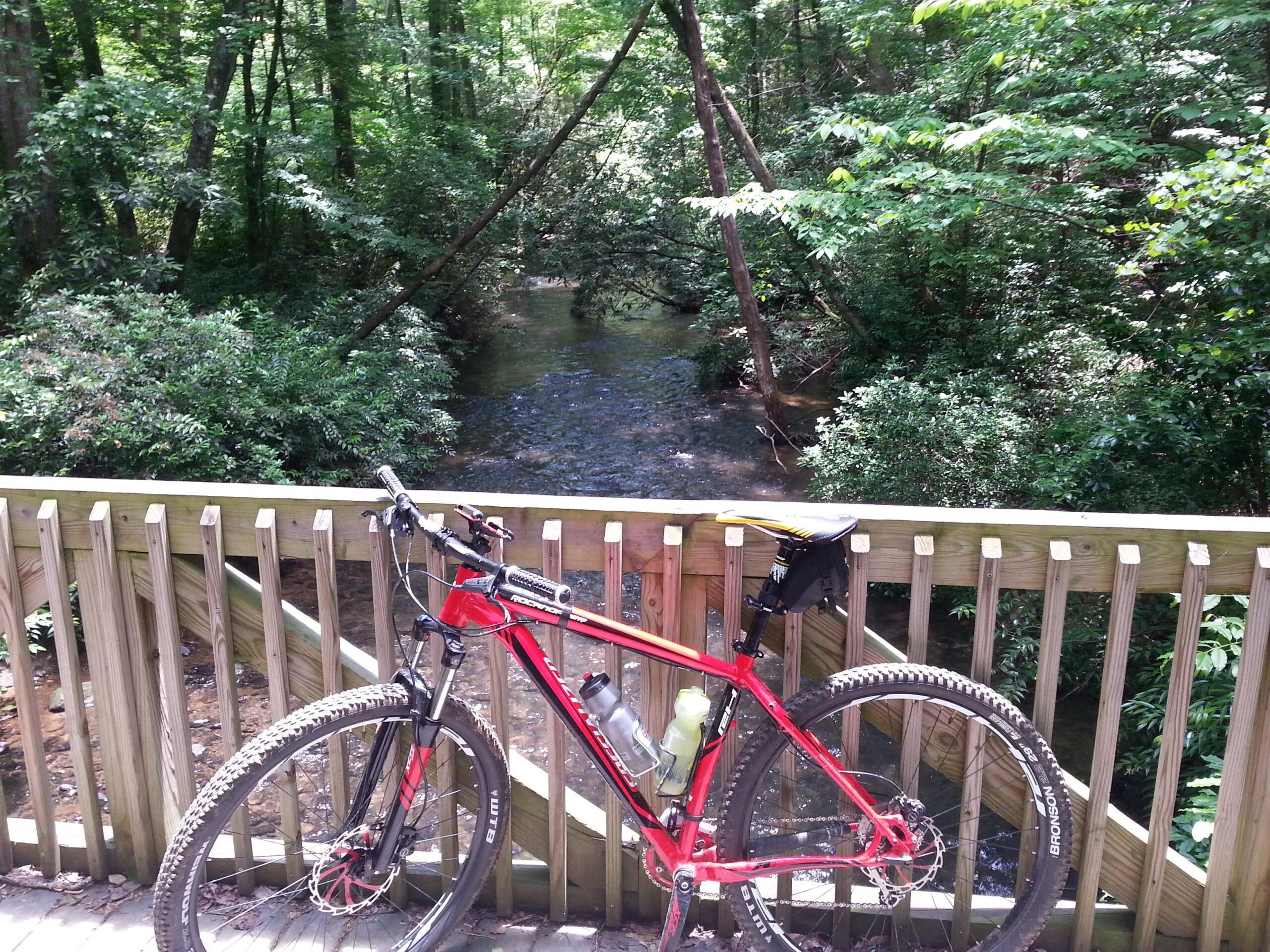 A red mountain bike is resting on a wooden bridge overlooking a serene stream surrounded by lush green vegetation and trees. Sunlight filters through the leaves, creating a peaceful outdoor scene. Unicoi State Park mountain bike trail.
