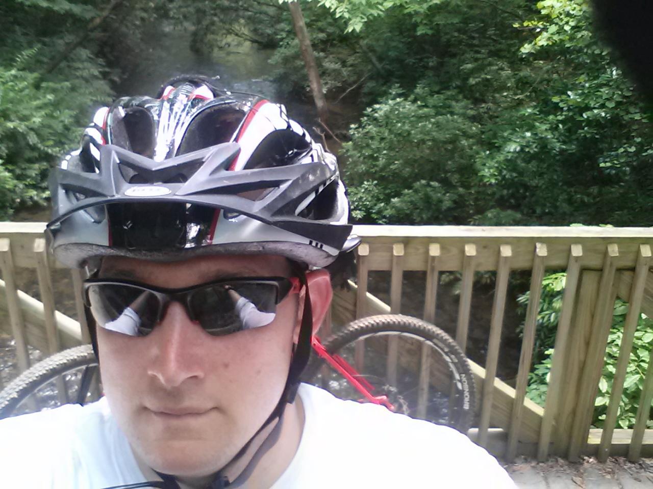 A person wearing a black and red bike helmet and sunglasses poses for a selfie on a wooden bridge. In the background, a creek can be seen surrounded by lush green foliage. A bicycle wheel is partially visible beside them. Unicoi State Park mountain bike trail.