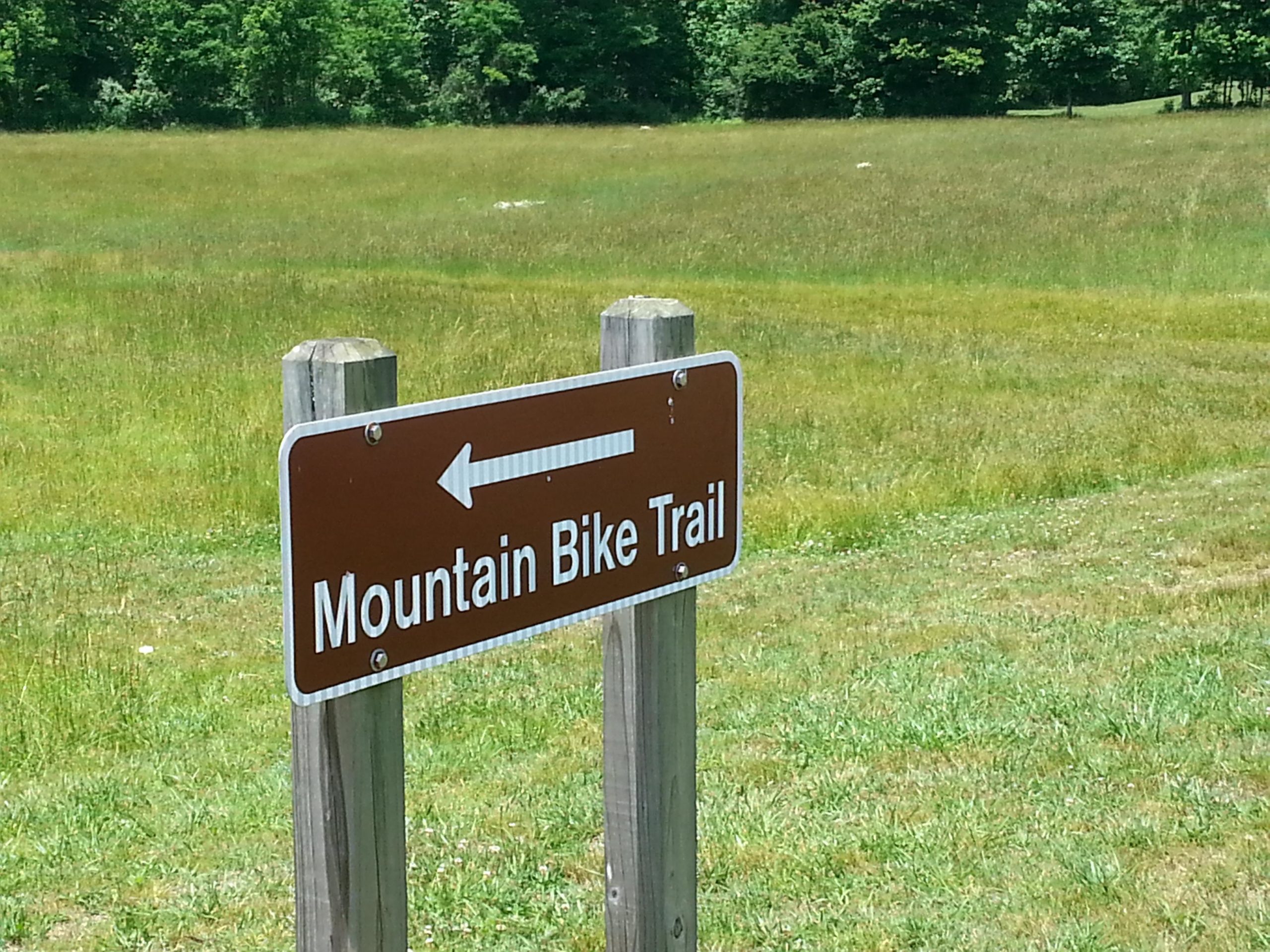 Sign indicating the direction of a mountain bike trail, with an arrow pointing to the left. The sign is mounted on a wooden post, surrounded by a grassy field and trees in the background. Unicoi State Park mountain bike trail.