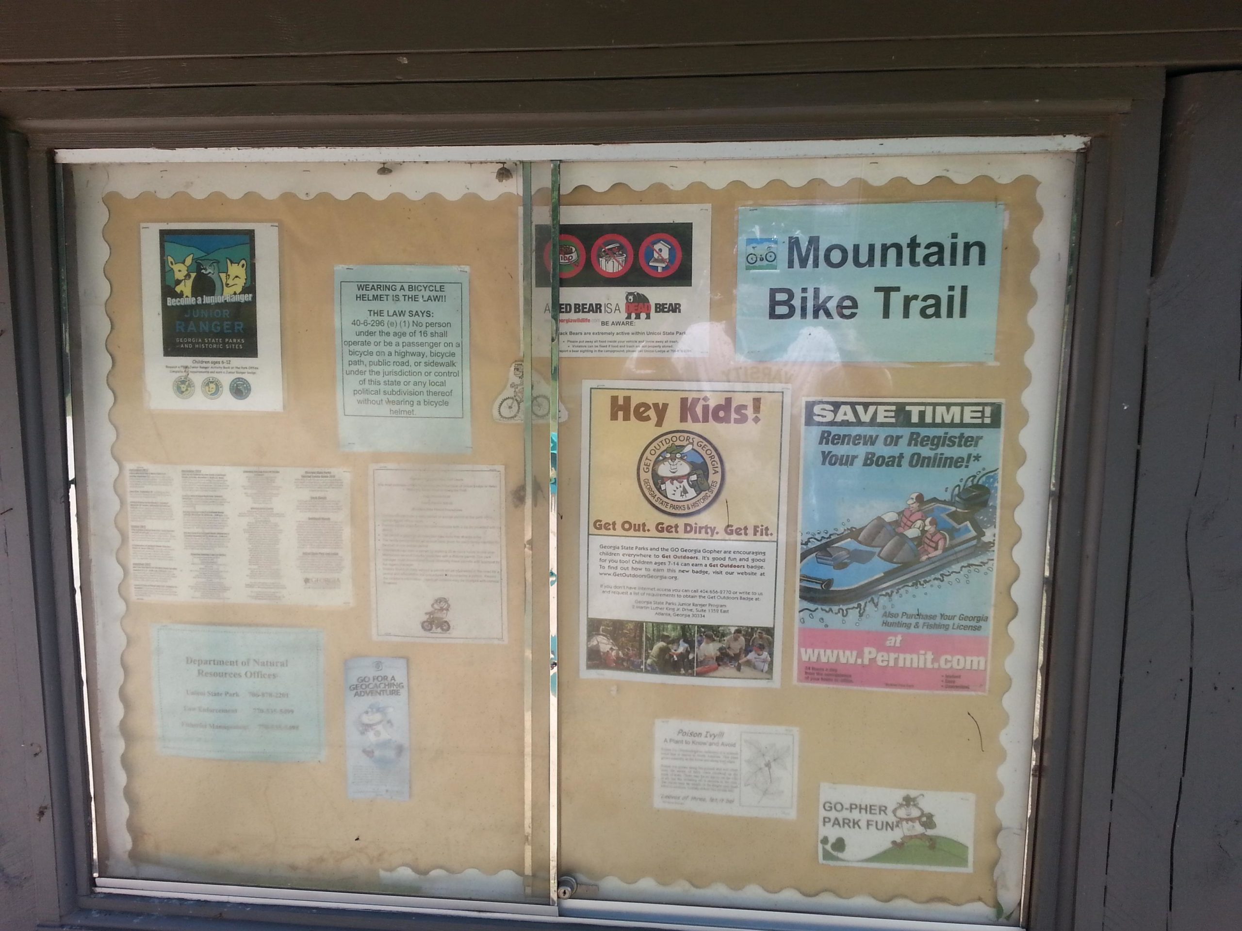A bulletin board display featuring various informational flyers and signs, including a "Mountain Bike Trail" notice, safety guidelines about wearing bicycle helmets, a Junior Ranger program advertisement, and promotional material for boating registration. The background is a brown paper with a scalloped border, and the board contains colorful graphics and text aimed at outdoor enthusiasts and families. Unicoi State Park mountain bike trail.