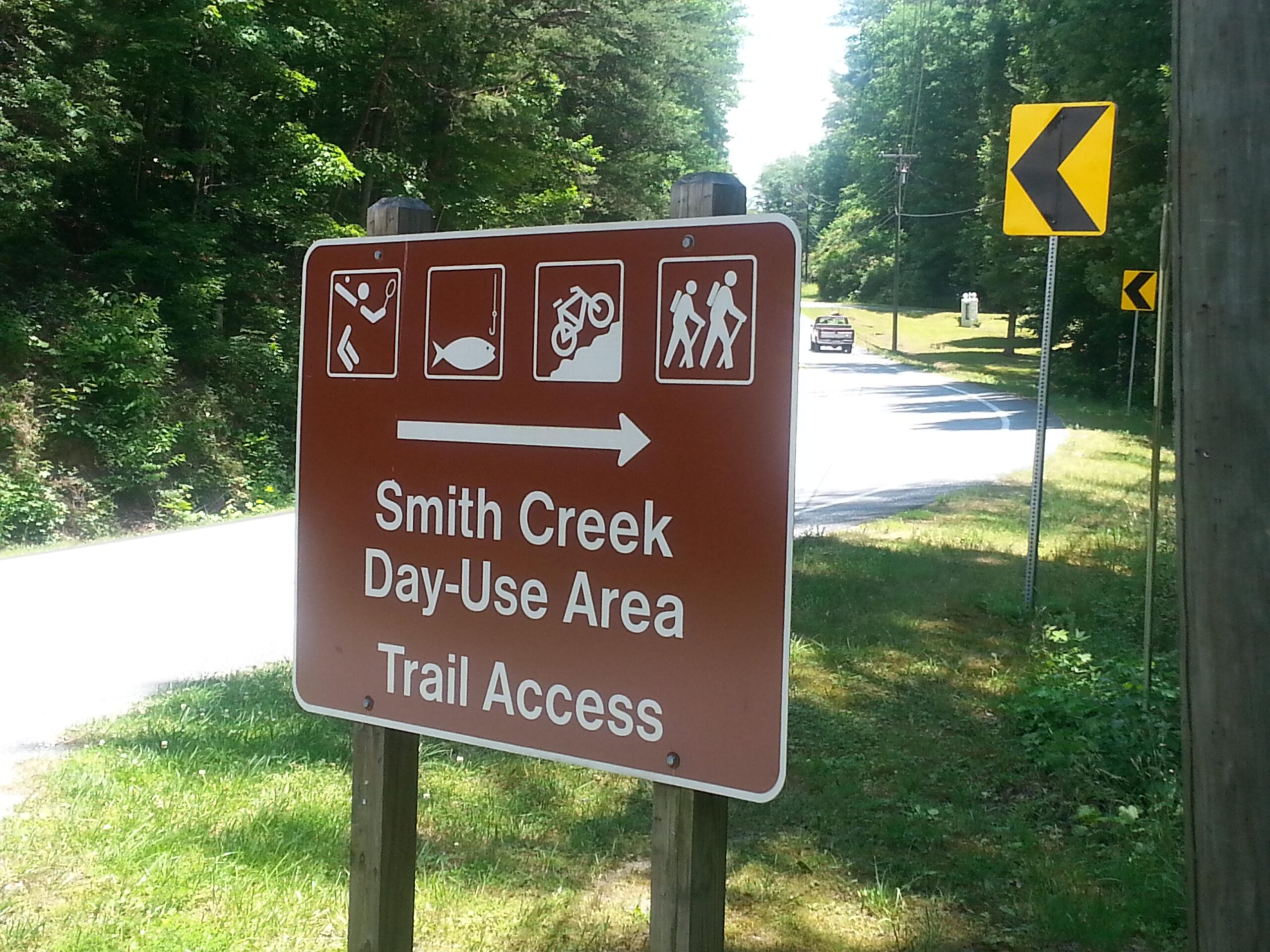 Sign for Smith Creek Day-Use Area, indicating trail access with icons for fishing, hiking, and biking. The sign points left, with a curve warning sign visible in the background, surrounded by greenery and a road. Unicoi State Park mountain bike trail.