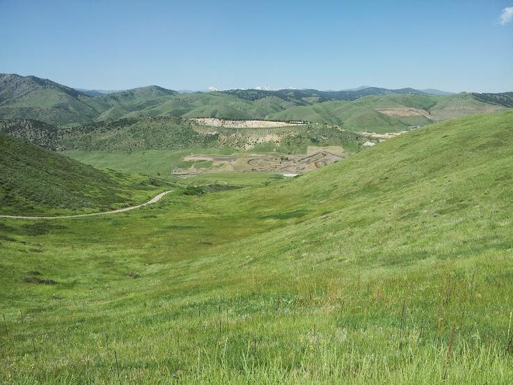 A panoramic view of lush green hills under a clear blue sky, with gentle slopes leading down to a distant valley where the outline of a construction site is visible. The landscape features rolling terrain with a dirt path winding through the grass. Green Mountain mountain bike trail.