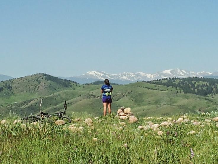 A person standing on a grassy hillside, looking out over a mountainous landscape with snow-capped peaks in the background. The foreground features rocks and wildflowers, while a bicycle is lying on the ground nearby. The sky is clear and blue, indicating a sunny day. Green Mountain mountain bike trail.