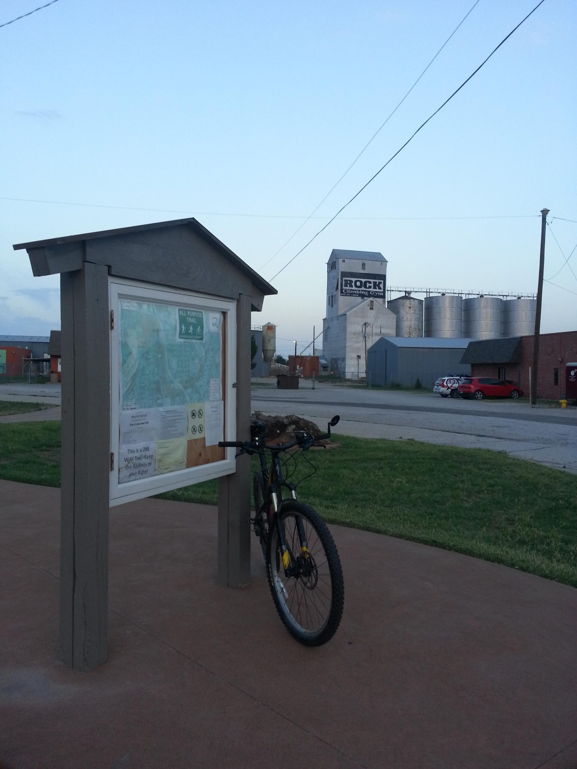 A bicycle leans against a wooden information board displaying a map and trail guidelines, set in an open area with grass and a concrete path. In the background, a large building with a sign that reads "ROCK" is visible, along with silos and other structures. The scene is captured during twilight, with a clear sky above. Wee-chi-ta mountain bike trail.