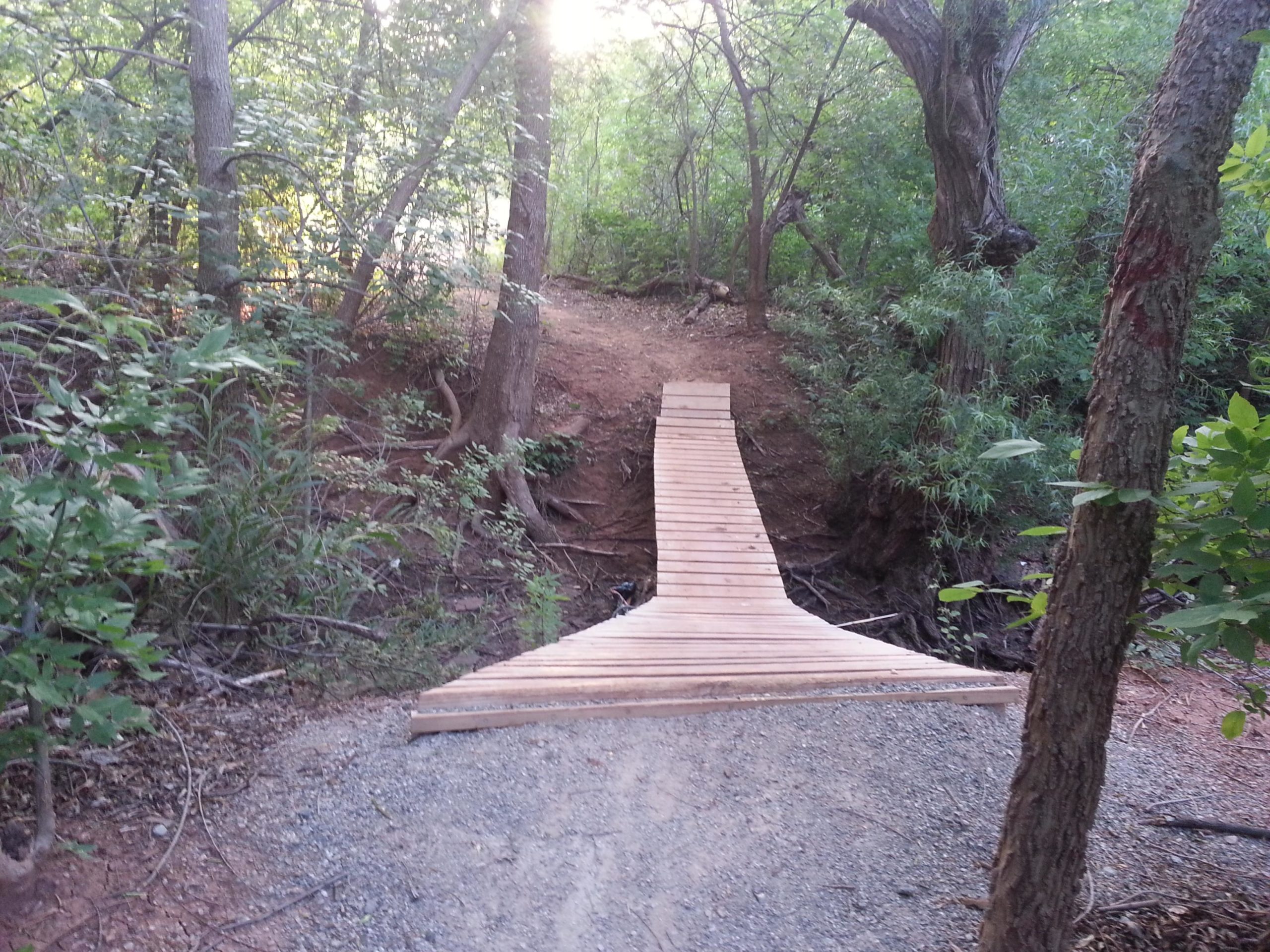 A wooden bridge spans a small gap in a wooded area, surrounded by lush green trees and foliage. The path leading to the bridge is made of gravel, and the sunlight filters through the trees, creating a serene and inviting atmosphere. Wee-chi-ta mountain bike trail.