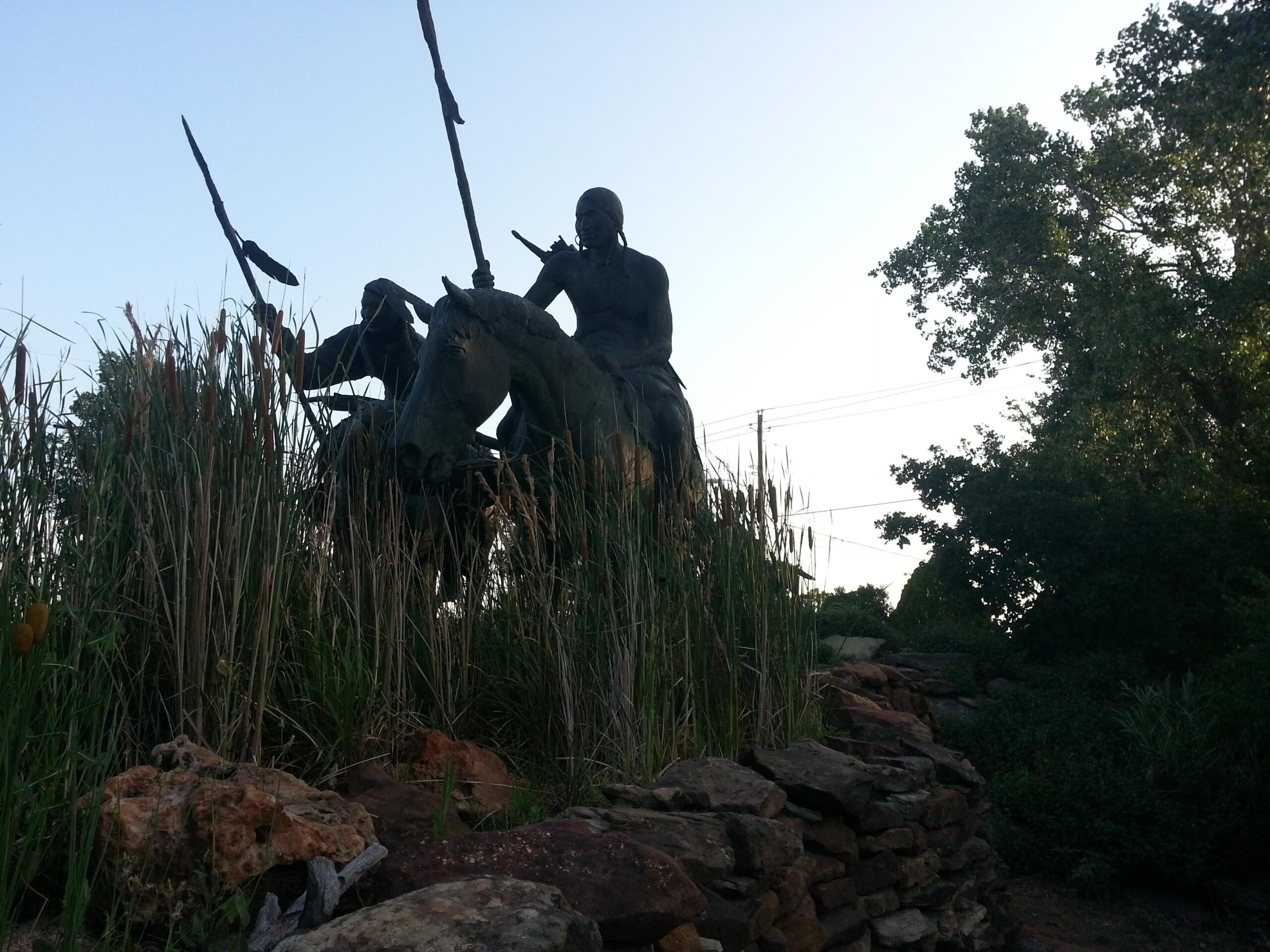 A silhouette of a large statue depicting warriors on horseback, holding spears, set against a backdrop of tall grasses and foliage. The scene is illuminated by soft evening light, with a clear sky above. A stone wall can be seen in the foreground, adding to the natural setting. Wee-chi-ta mountain bike trail.