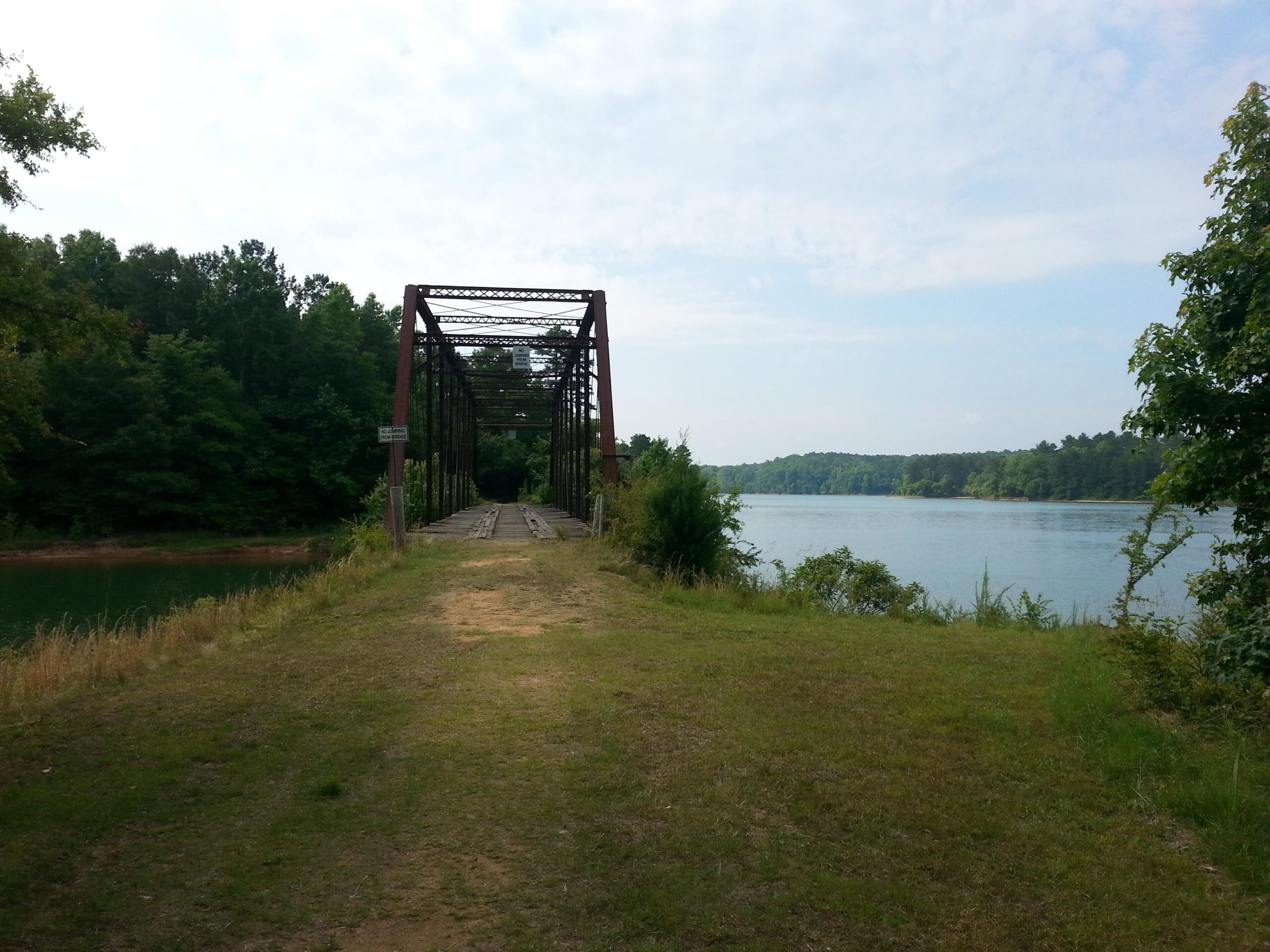 A rusty, old bridge extends over a calm river, surrounded by lush greenery. The path leading to the bridge is grassy and well worn, with trees lining the bank. The sky is partly cloudy, adding a serene ambiance to the landscape. Cottage Loop Trail mountain bike trail.