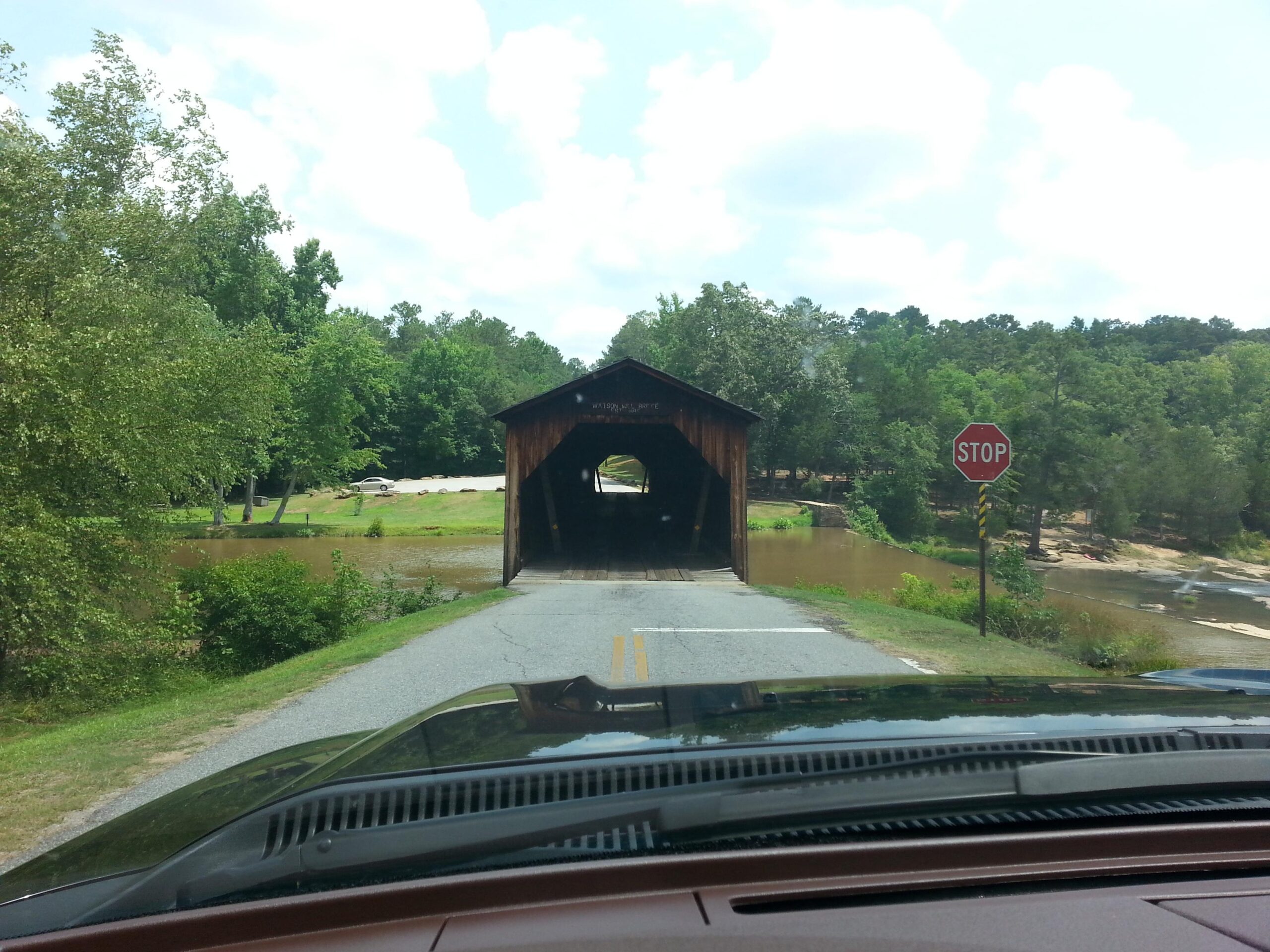 A scenic view of a covered bridge in the foreground, surrounded by lush greenery and a calm, flooded river. A stop sign is visible on the right side of the road, and a vehicle can be seen near the bridge entrance. The sky is partly cloudy, adding to the tranquil atmosphere. Watson Mill Bridge State Park mountain bike trail.