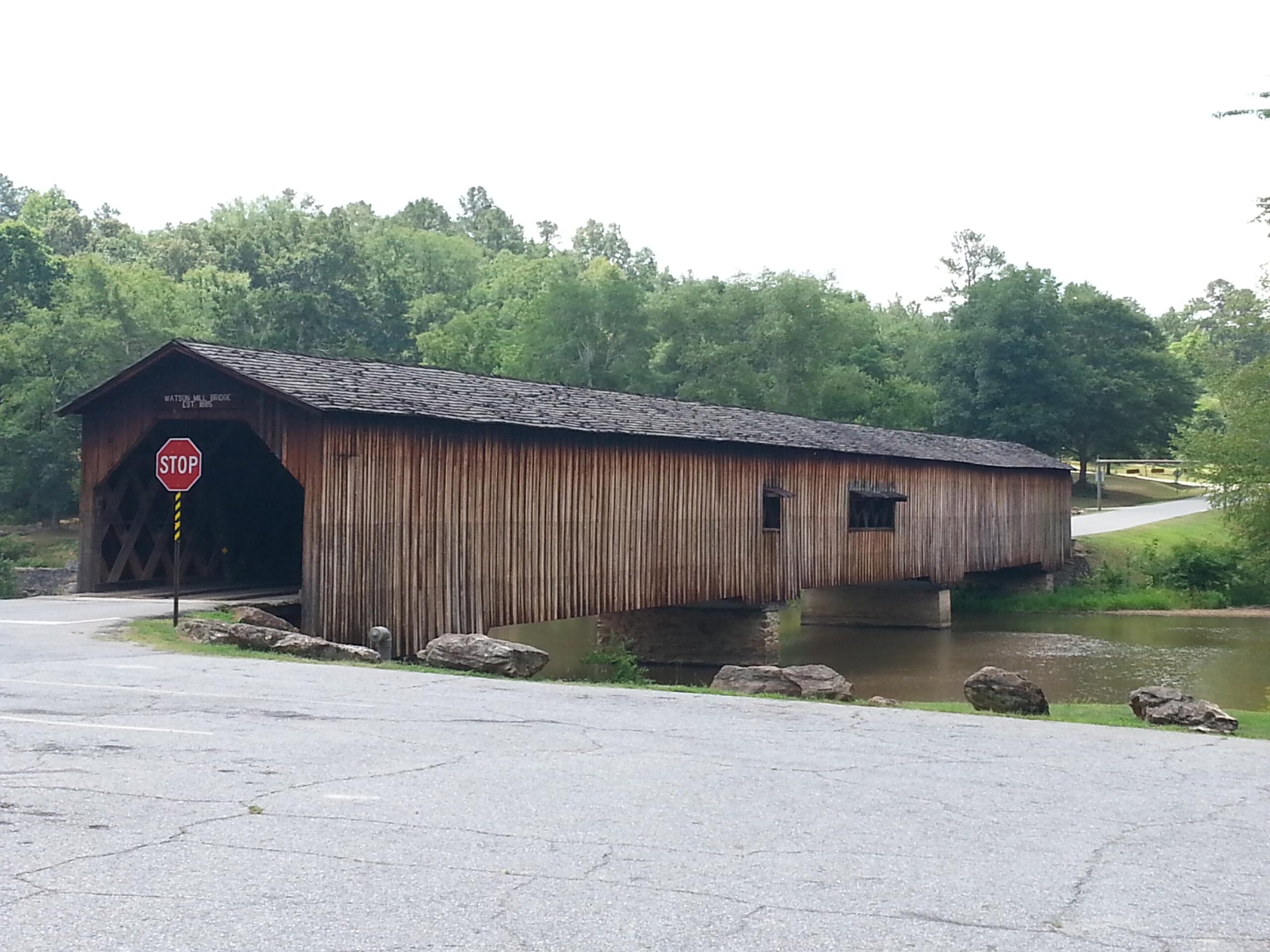 A wooden covered bridge spanning a small body of water, with a stop sign at the entrance and a scenic backdrop of lush green trees. The bridge features a rustic design, with a peaked roof and visible wooden beams, situated near a winding road and rocky shoreline. Watson Mill Bridge State Park mountain bike trail.