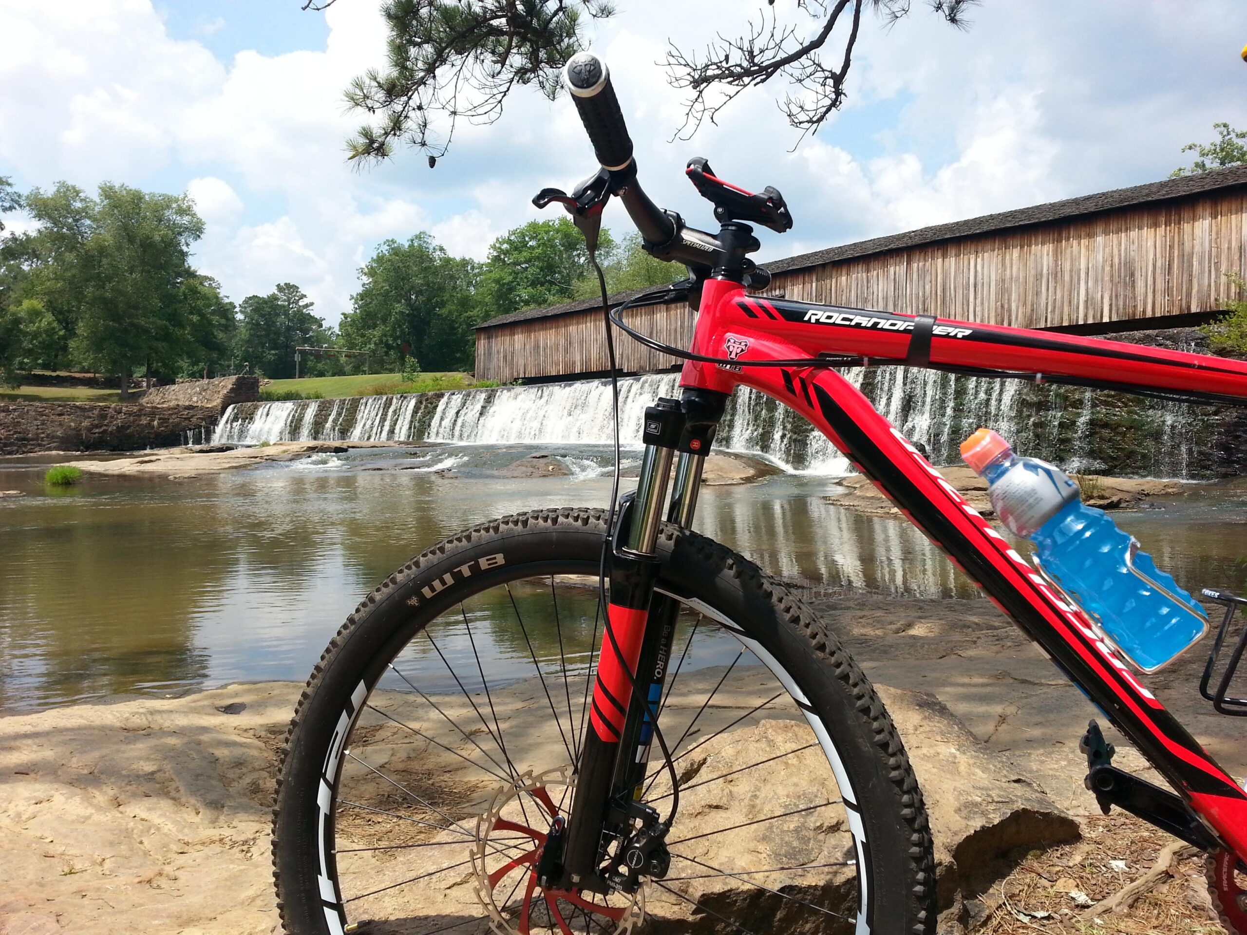 A close-up view of a red mountain bike leaning against a rock near a waterfall. The bike has a water bottle in a holder, and the scene features lush green trees and a blue sky with fluffy white clouds in the background. Watson Mill Bridge State Park mountain bike trail.