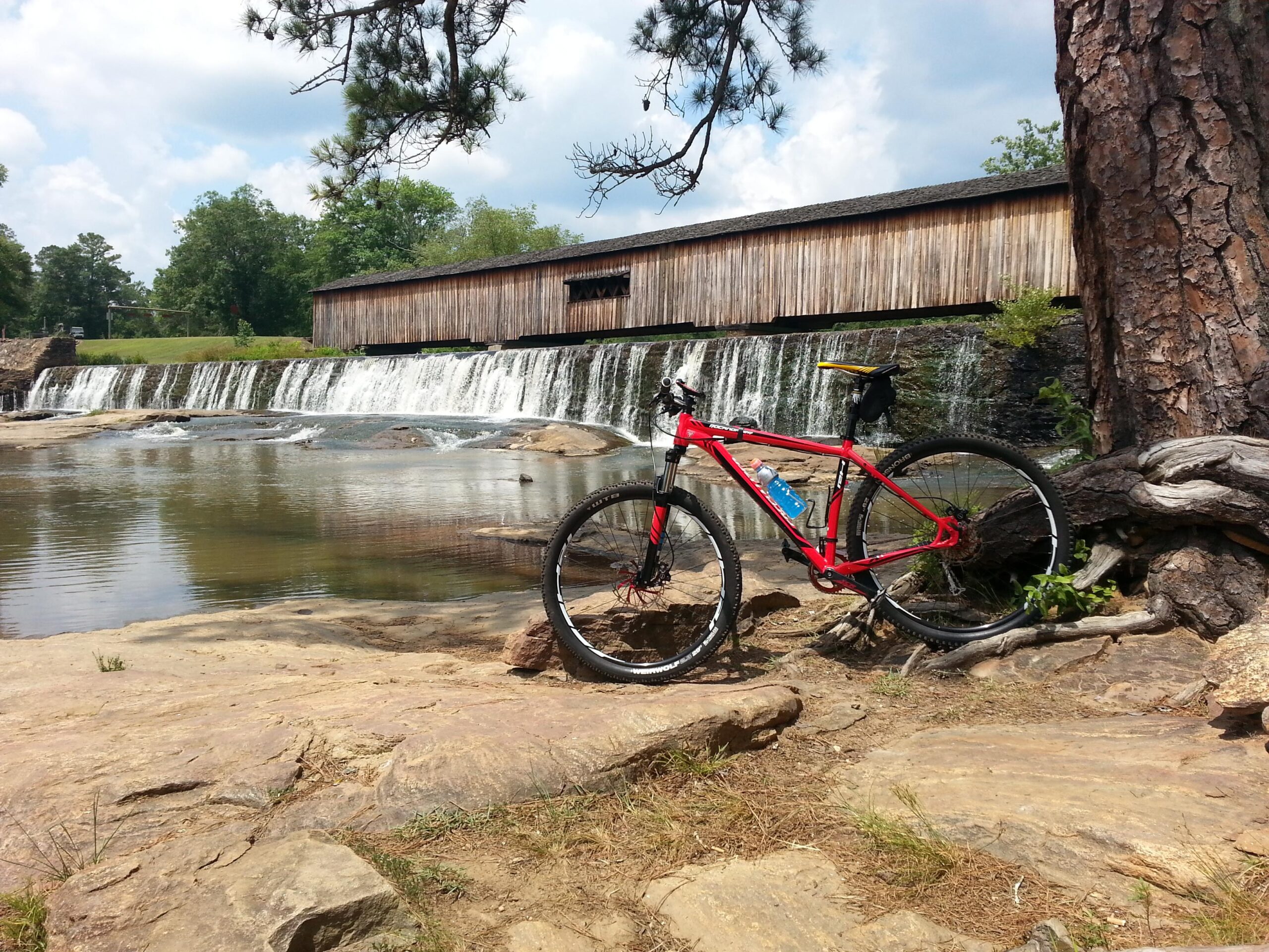 A red mountain bike rests on rocky terrain beside a calm body of water, with a small waterfall cascading in the background. A covered wooden structure is visible across the water, surrounded by green trees and a partly cloudy sky. Watson Mill Bridge State Park mountain bike trail.