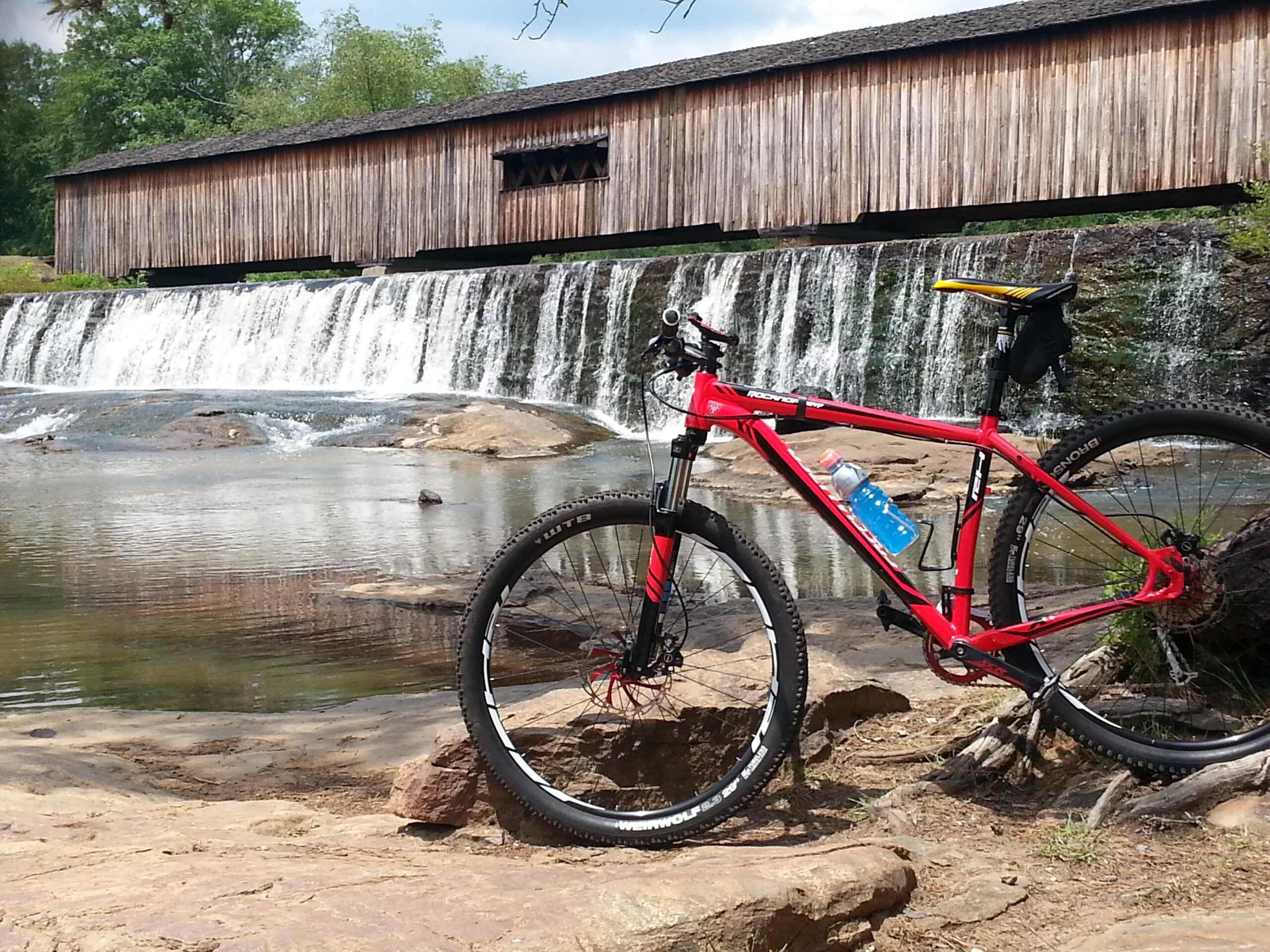 A red mountain bike is resting on a rocky shore near a tranquil river, with a waterfall cascading in the background. A wooden covered bridge is visible above the waterfall, surrounded by lush greenery under a partly cloudy sky. A water bottle is attached to the bike frame. Watson Mill Bridge State Park mountain bike trail.