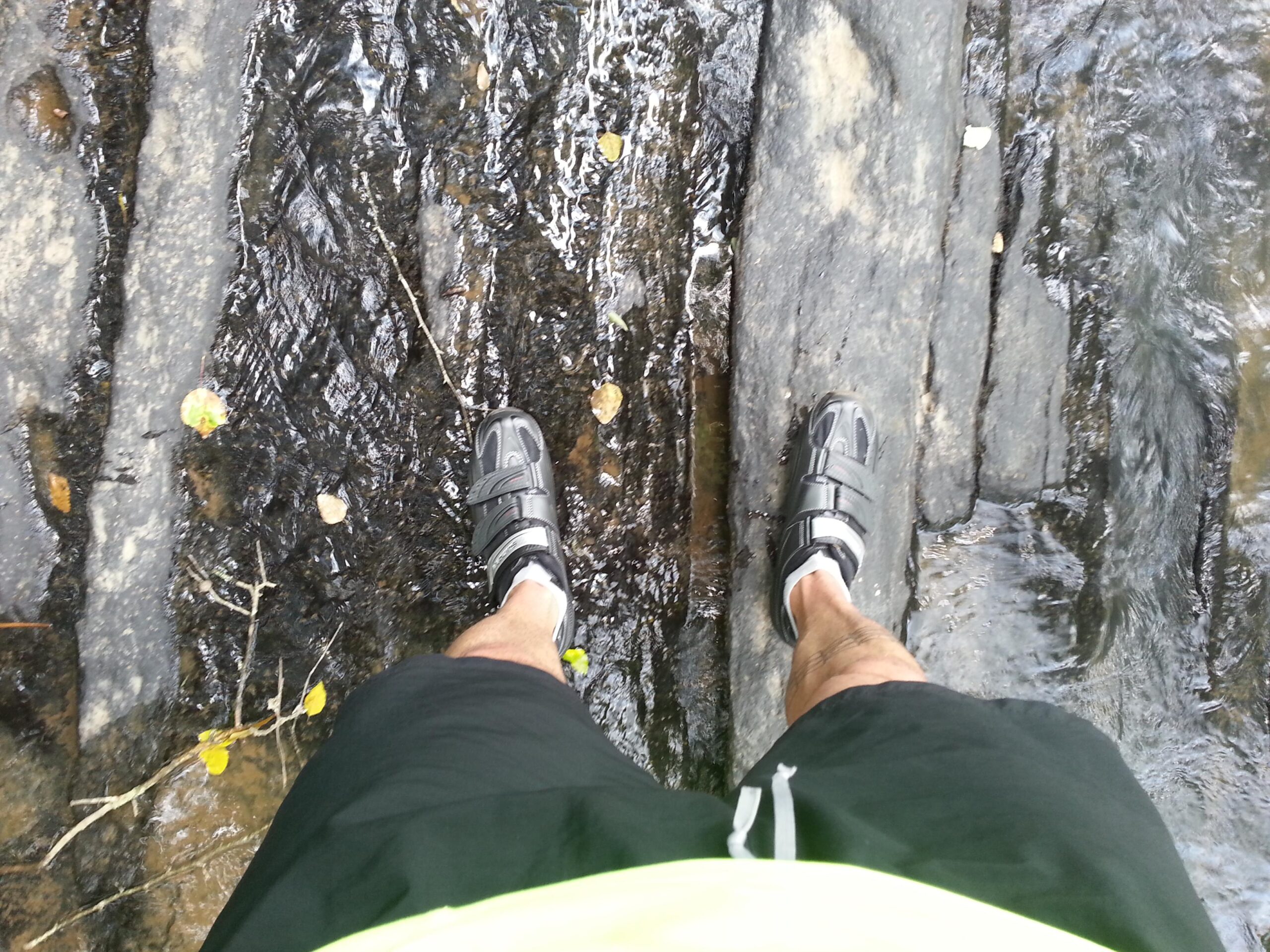 A top-down view of a person's legs standing on a rocky surface beside flowing water. The individual is wearing black shorts and sporty shoes with straps. Leaves are scattered around on the wet rocks, and the water reflects some of the rocks' textures. Watson Mill Bridge State Park mountain bike trail.