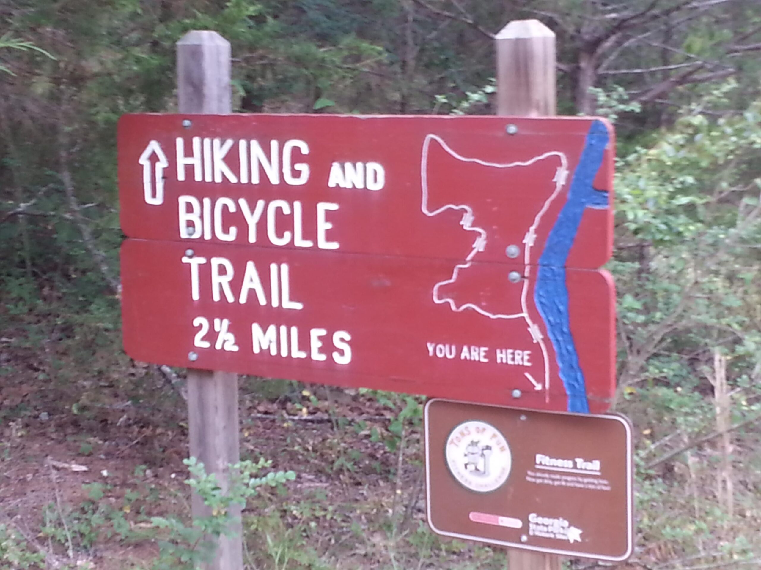 Signpost indicating a hiking and bicycle trail, featuring an arrow pointing up, the text "Hiking and Bicycle Trail," a distance of "2½ miles," and a map indicating a location marked "You Are Here," set against a natural background with greenery. Watson Mill Bridge State Park mountain bike trail.