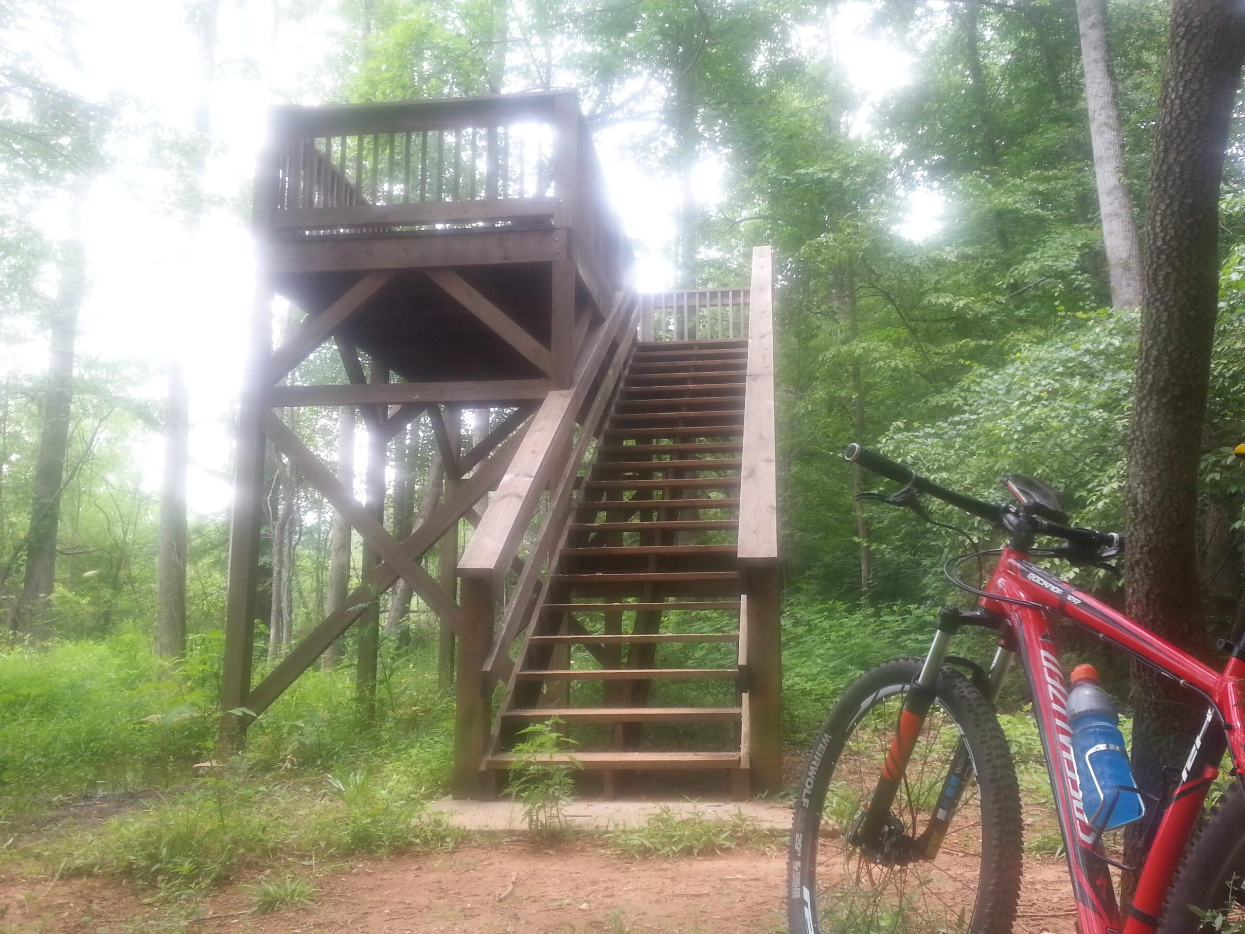 A wooden observation deck with stairs surrounded by lush greenery, alongside a mountain bike with a water bottle attached, set in a forested area. Victoria Bryant State Park mountain bike trail.