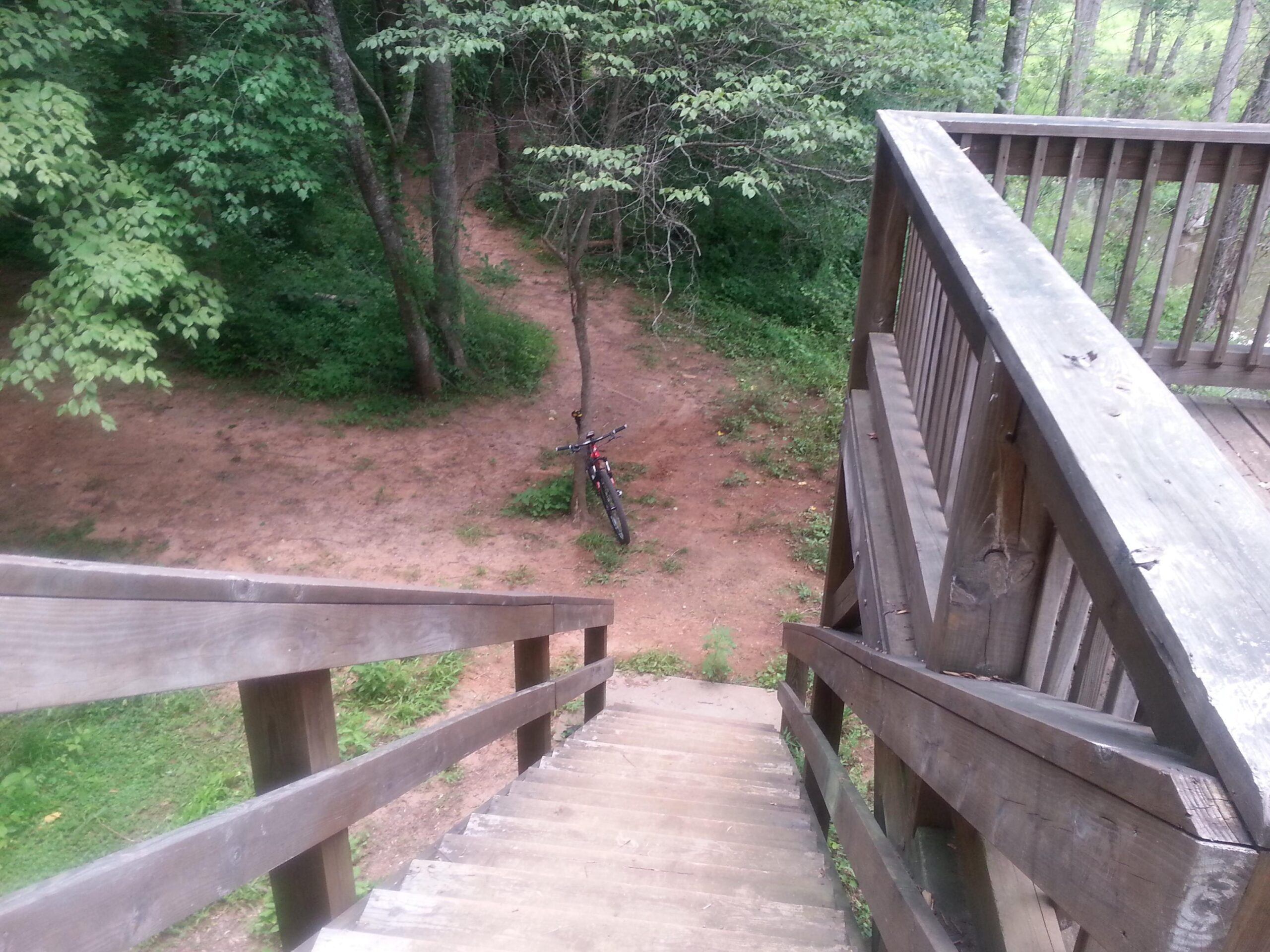 A wooden staircase leading down into a wooded area, with a bicycle parked on a dirt path below. The scene is framed by lush green trees and vegetation, creating a serene outdoor atmosphere. Victoria Bryant State Park mountain bike trail.