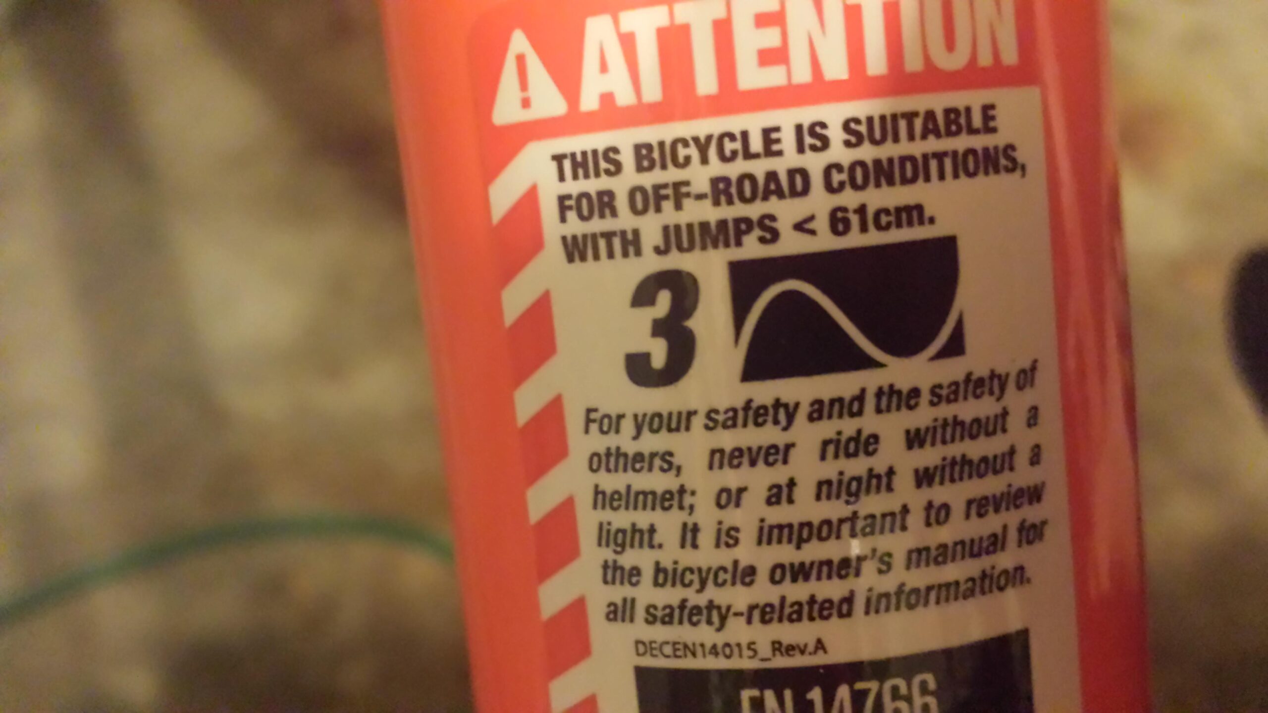 Motobecane FB4 Comp: Label on a bicycle indicating it is suitable for off-road conditions and jumps under 61 cm. Safety warnings advise wearing a helmet and having a light for nighttime riding. Additionally, it encourages reviewing the owner's manual for more safety information.
