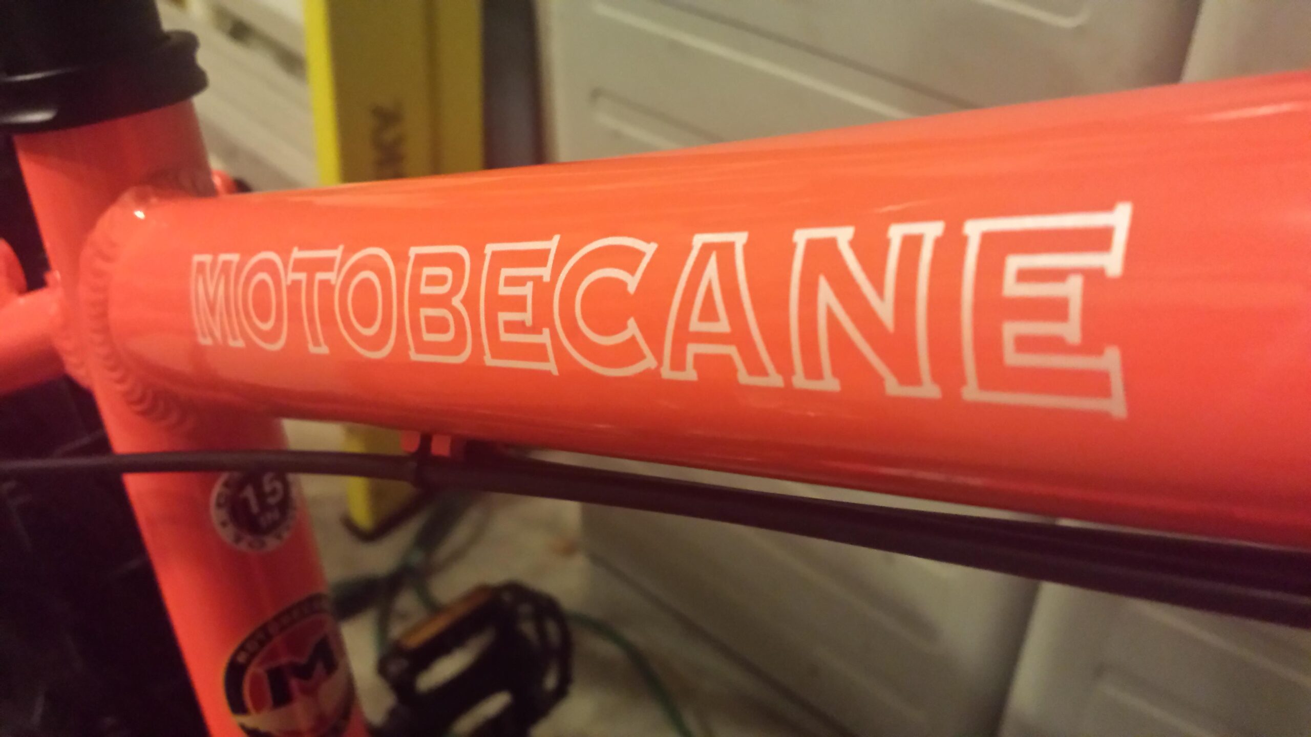 Motobecane FB4 Comp: Close-up image of a bright orange bicycle frame featuring the logo "MOTOBECANE" prominently displayed on the top tube. The background shows a blurred garage setting with tools and storage items.
