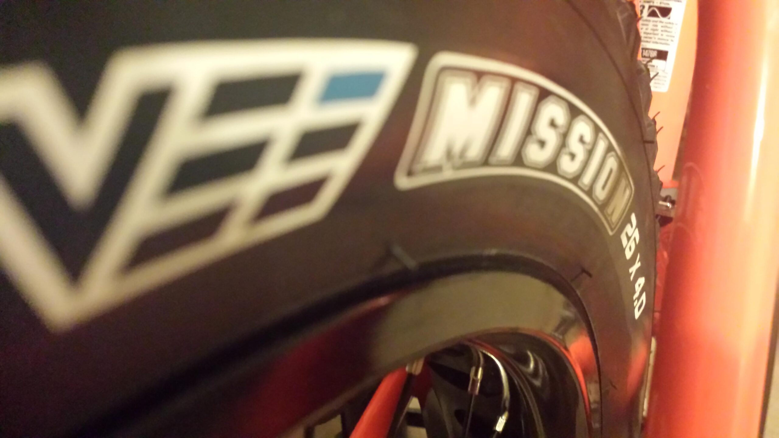 Motobecane FB4 Comp: Close-up image of a bicycle tire featuring the "Mission" logo, highlighting the tire's size specifications: "26 x 4.0." The background includes part of the bicycle frame in a reddish hue.