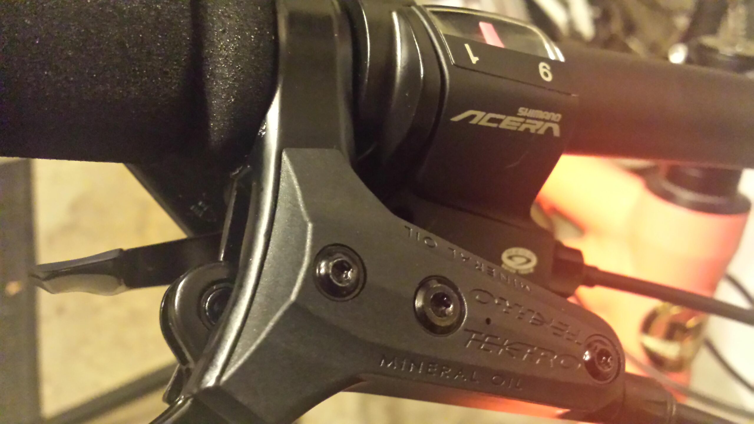 Motobecane FB4 Comp: Close-up of a bicycle brake lever and gear shifter, featuring Shimano Acera branding. The image shows the components in detail, including the lever mechanism and adjustment screws. The background is slightly blurred, emphasizing the equipment.