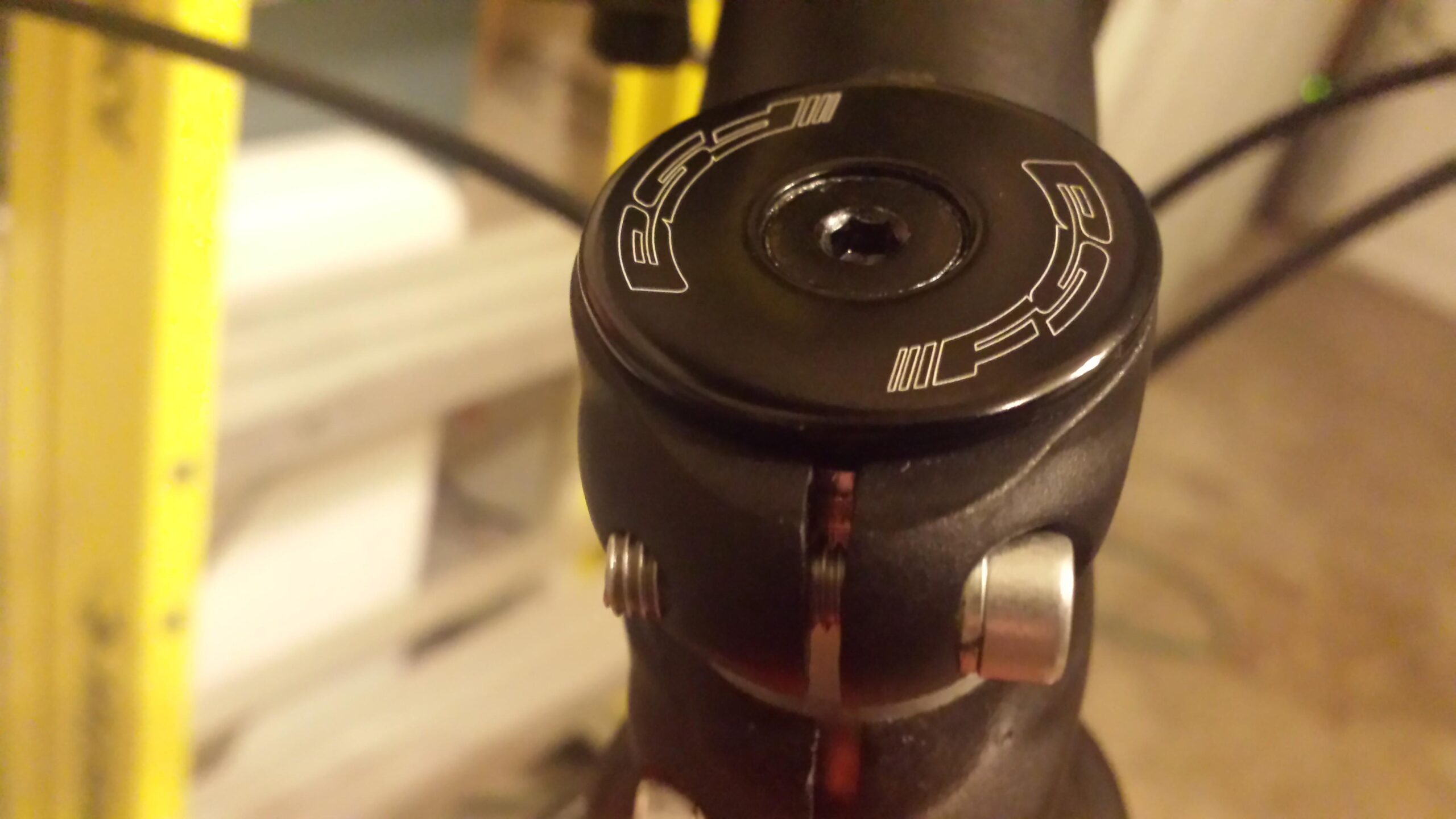 Motobecane FB4 Comp: Close-up image of a bicycle's headset with a black top cap featuring a logo, mounted on a black threaded stem. The background shows a blurred yellow object, likely a ladder or frame, indicating a workspace.